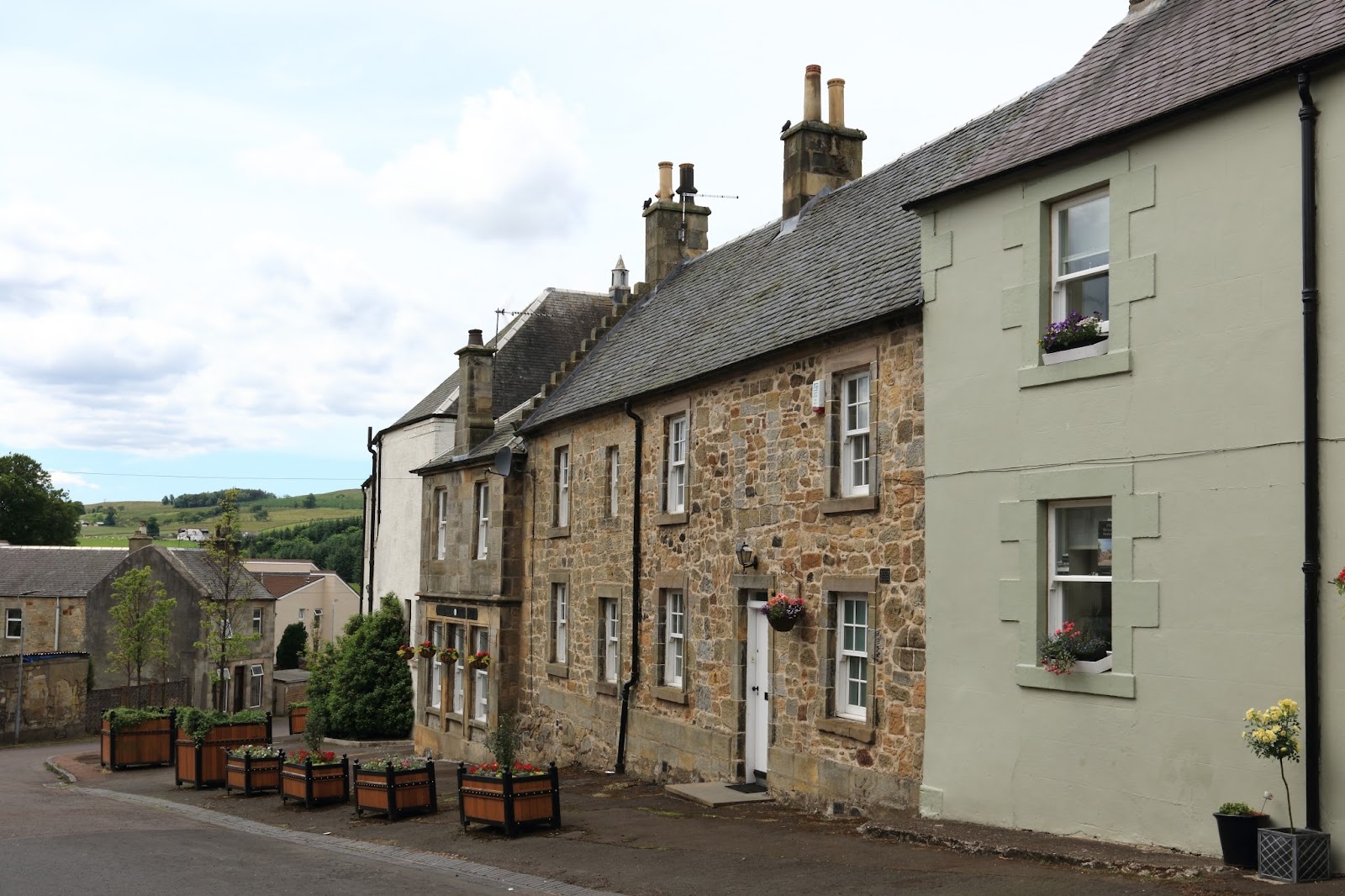 A row of stone cottages and a pale painted house along a quiet street in Douglas, flower tubs by the doors.
