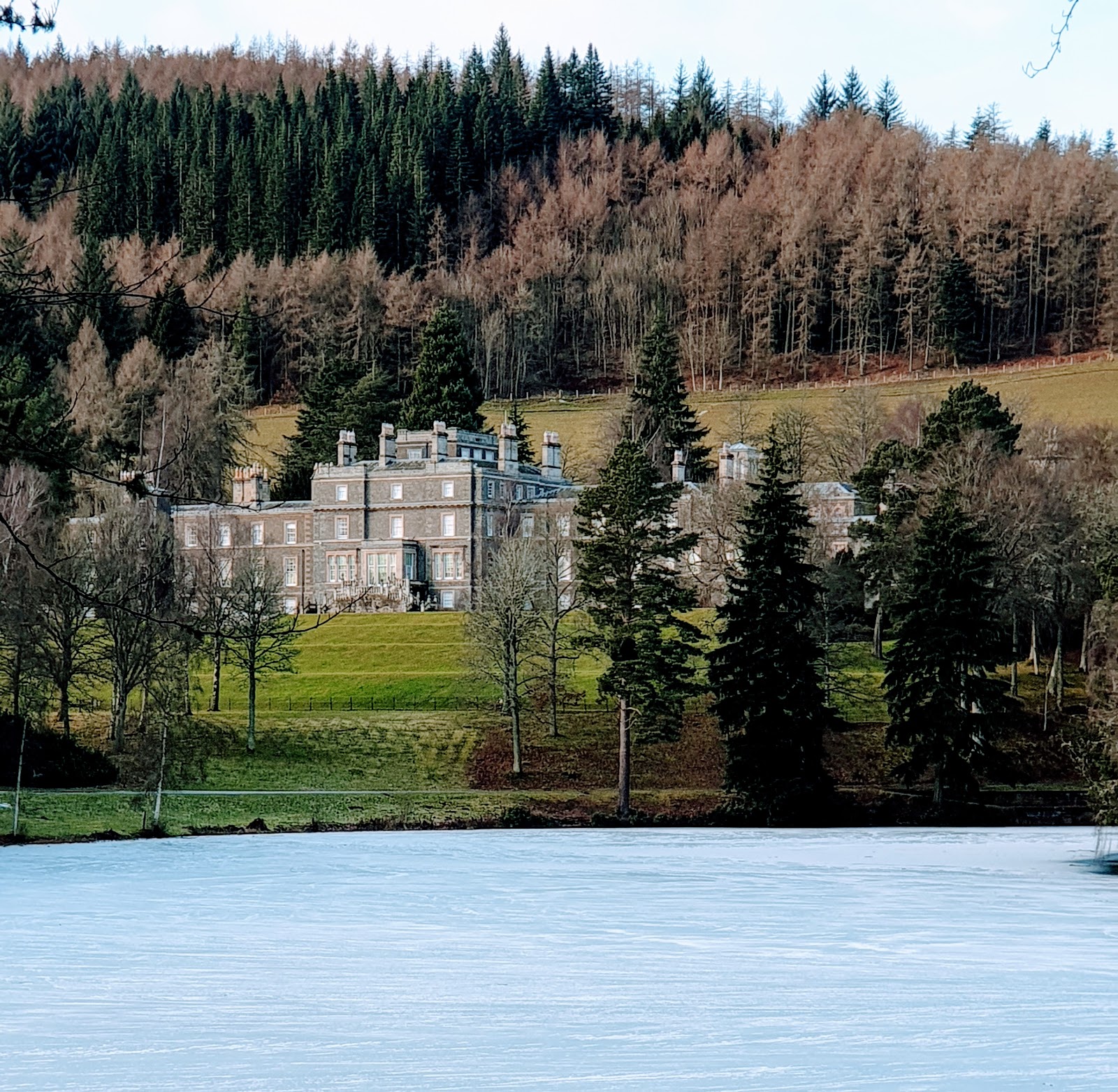 Bowhill House, the long pale mansion among trees on a hillside above a frozen loch, conifer woods behind.