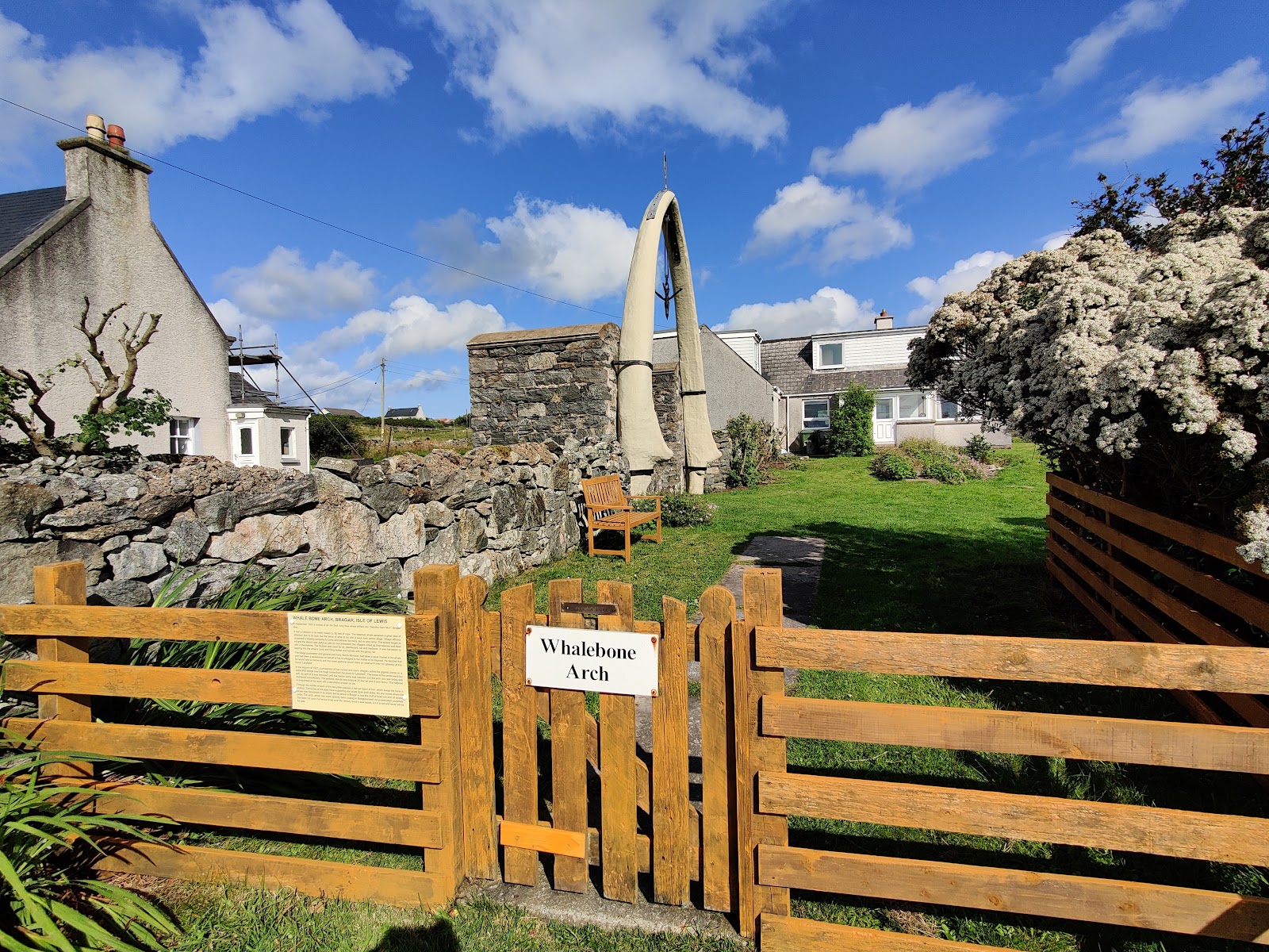 The Whalebone Arch in a garden at Bragar behind a wooden gate, cottages and a stone wall around it.
