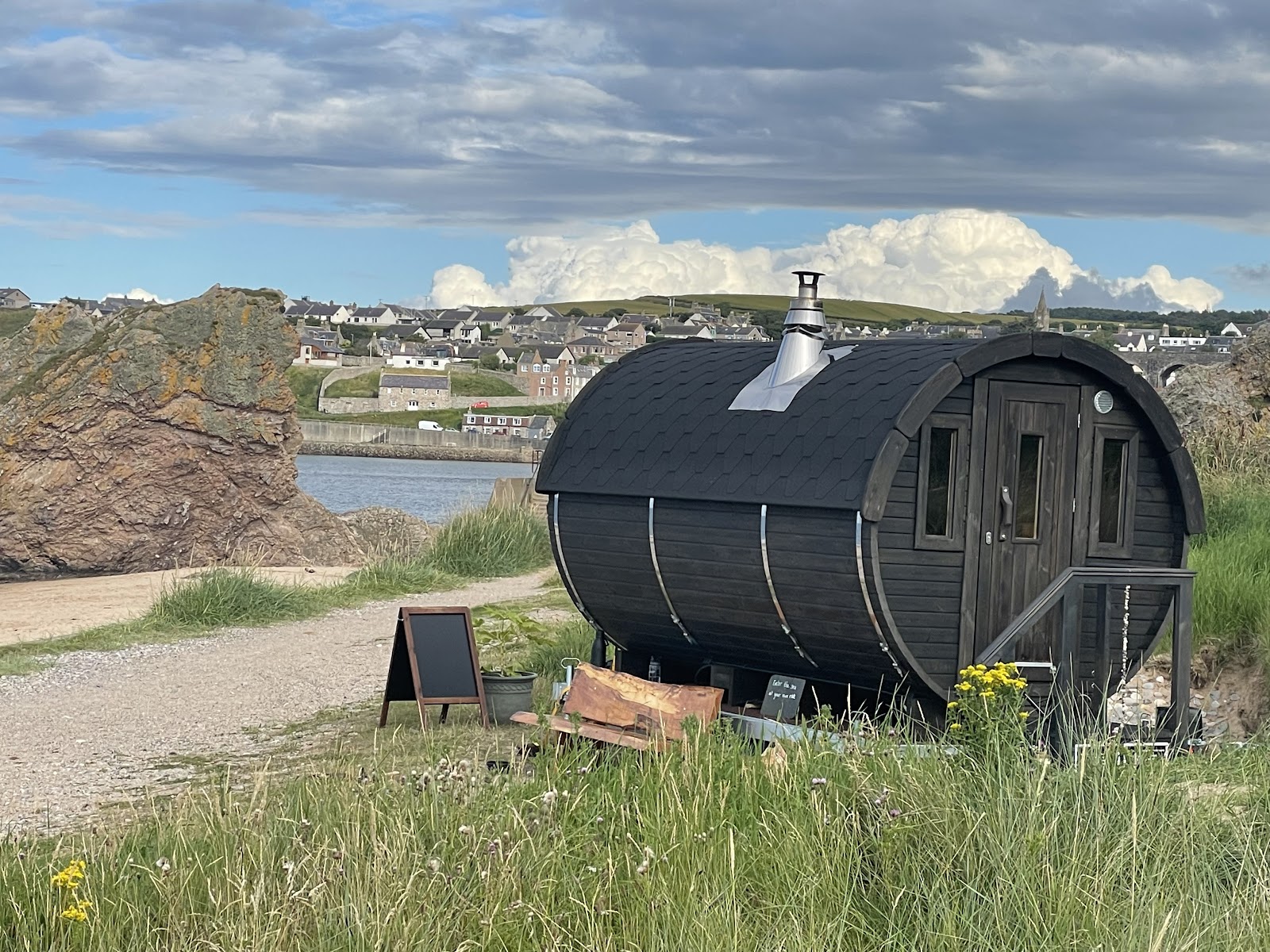 The black wood-fired barrel sauna on the dunes above Cullen beach, with the town and the Three Kings stacks behind.