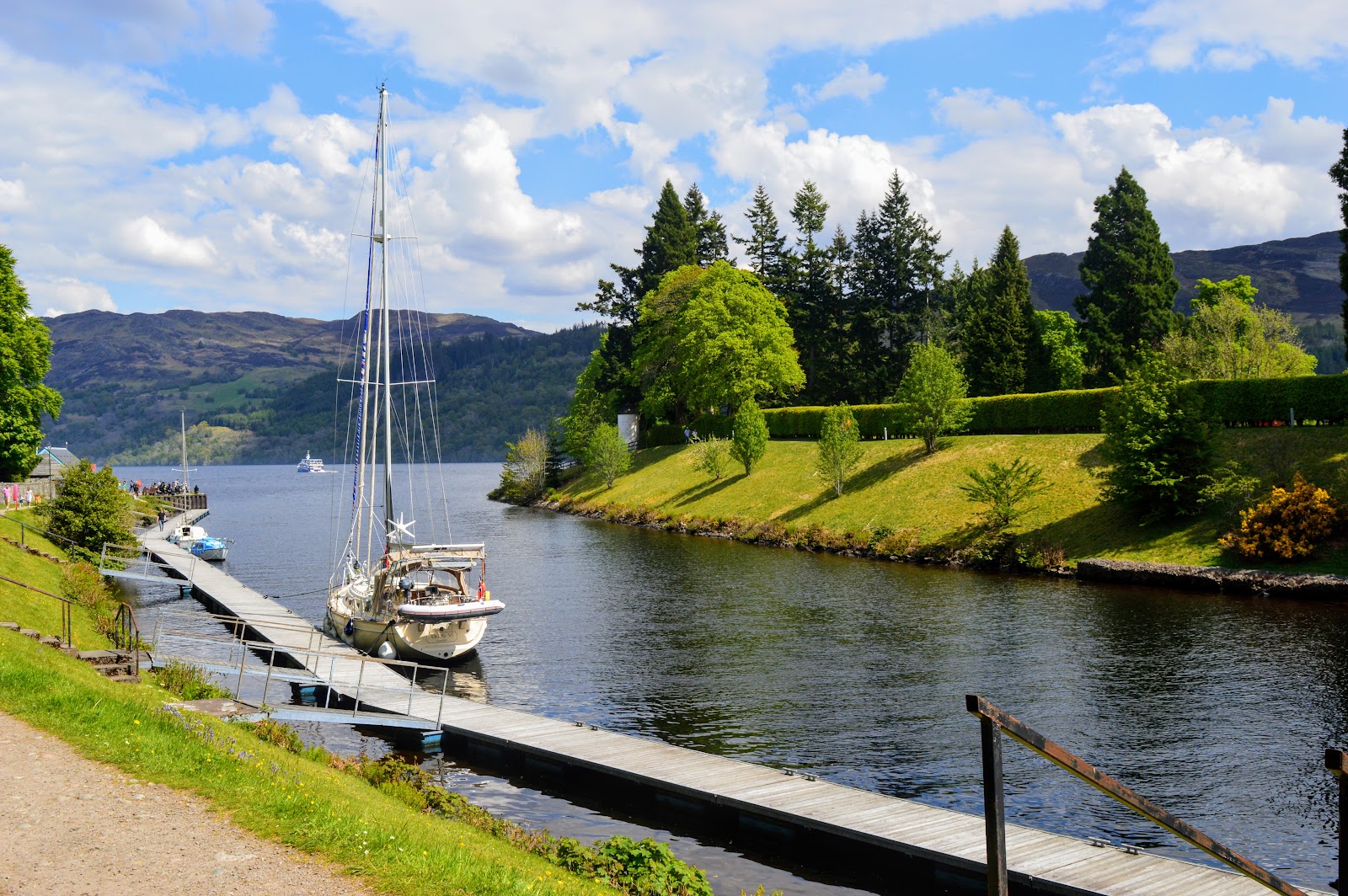 A yacht moored on a pontoon where the Caledonian Canal meets Loch Ness at Fort Augustus, trees and hills beyond.