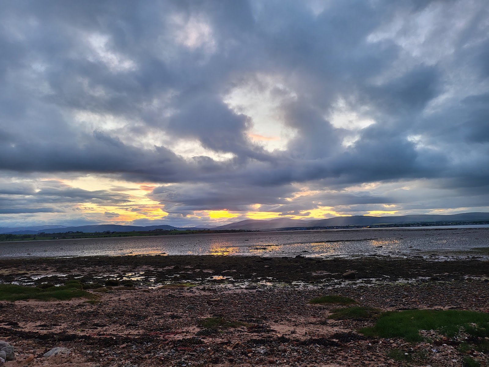 Sunset breaks through clouds over a mudflat estuary and distant hills