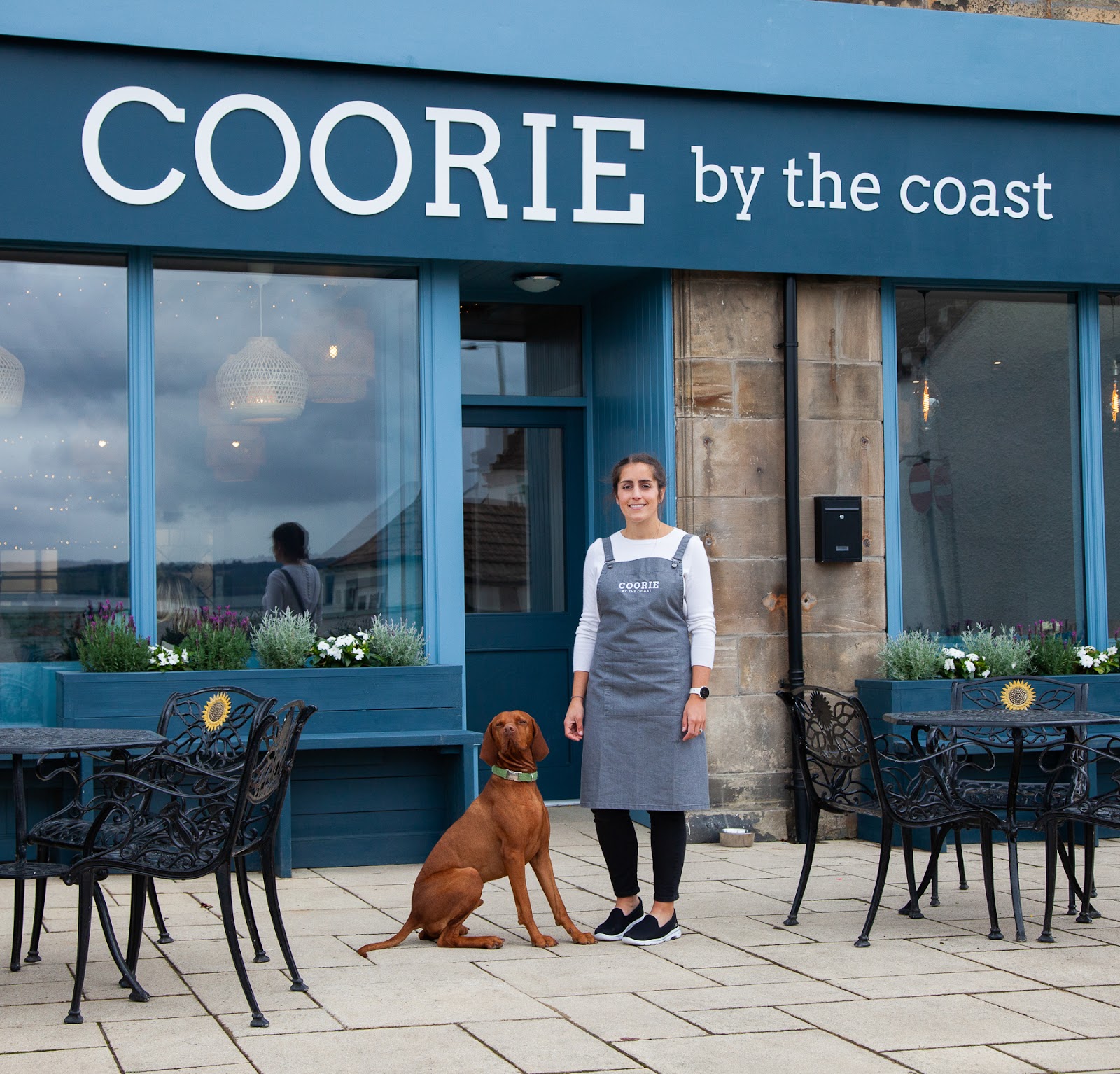 Aproned woman and dog stand outside Coorie by the Coast café