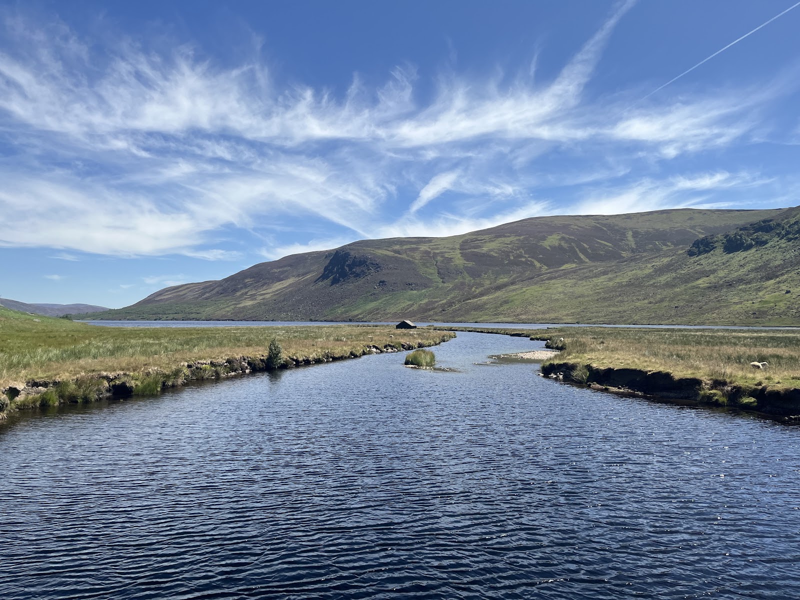 A river winds through grassy moorland towards a hillside loch and small hut