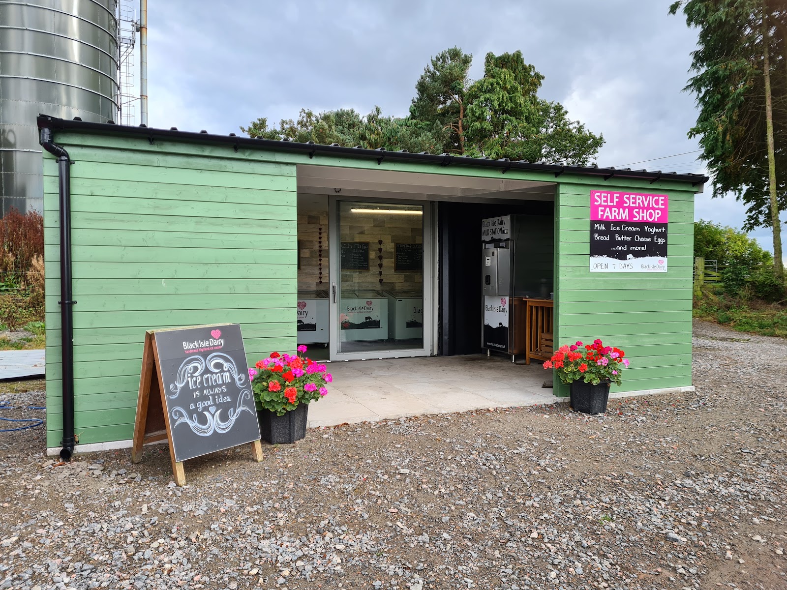 The Black Isle Dairy farm shop, a green timber building with a chalkboard and flower tubs on the gravel.