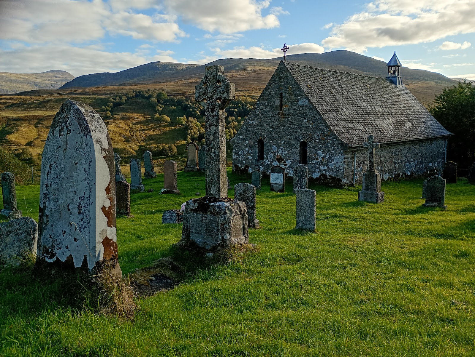 Cille Choirill, a small grey stone church with a bellcote among old gravestones and Celtic crosses on a green hillside, mountains beyond.