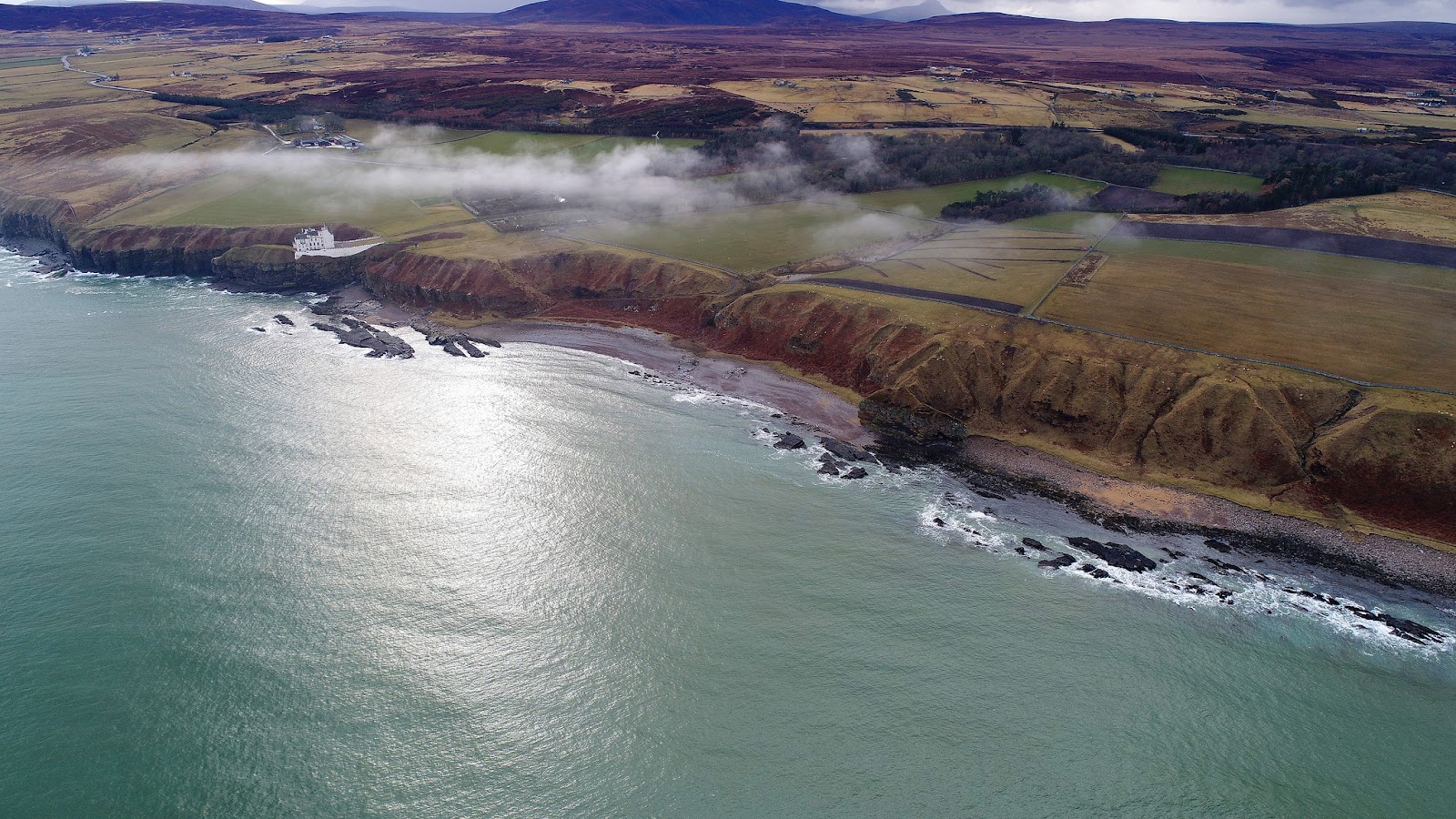 Aerial view of a lighthouse on cliffs above a rocky coastline, low cloud drifting over farmland