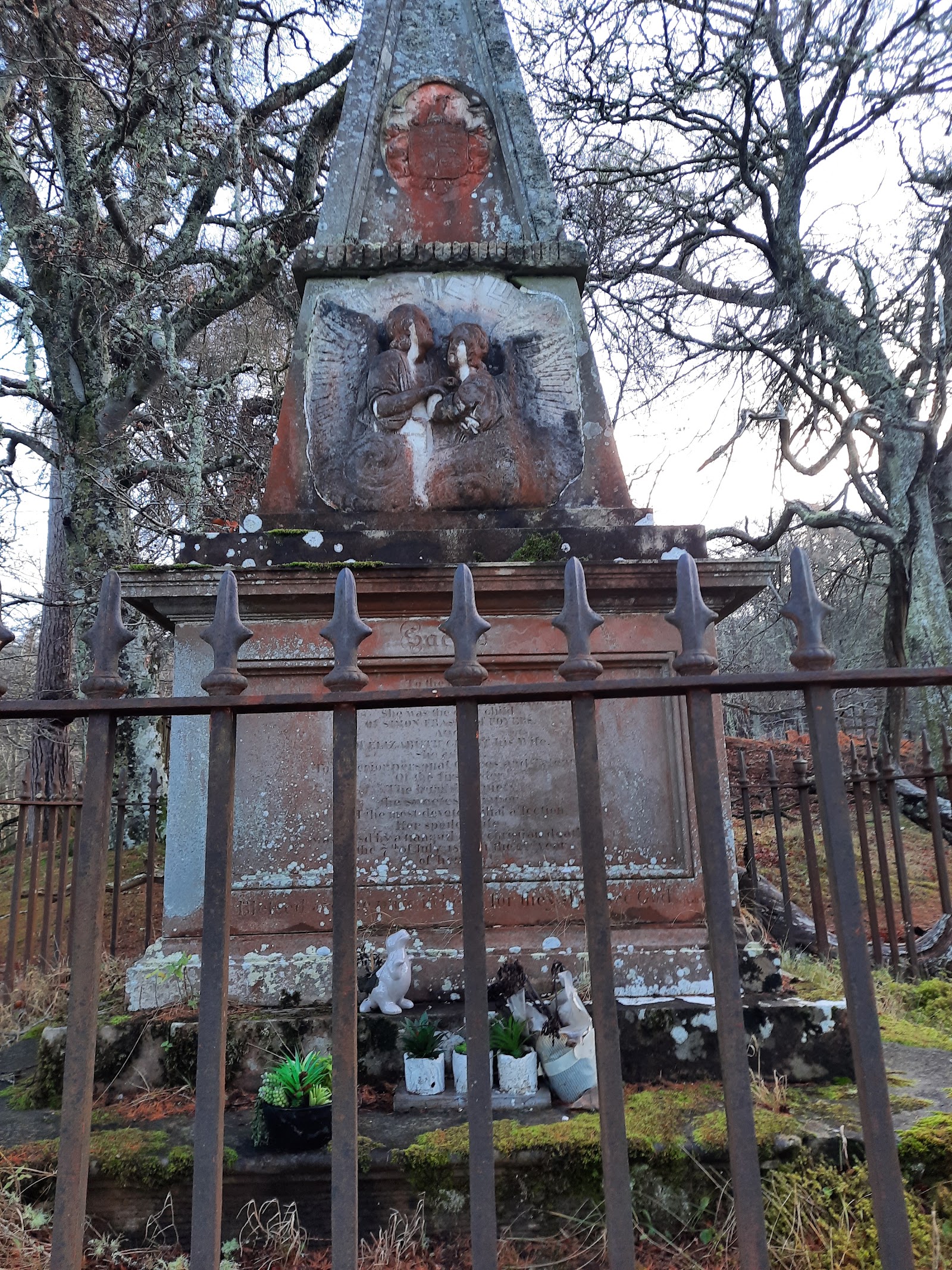 A red sandstone monument behind iron railings among bare trees in Boleskine burial ground.