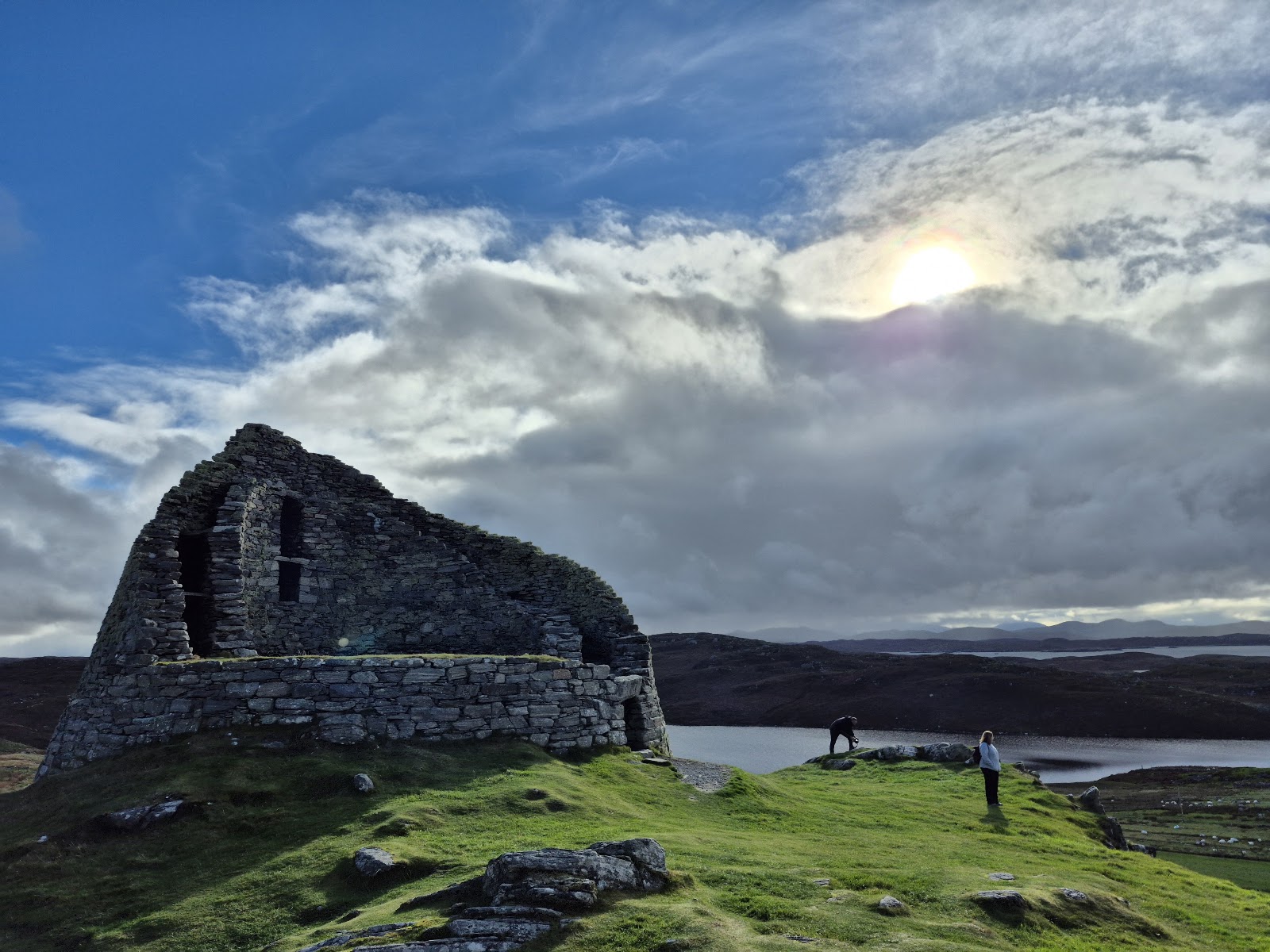 Dun Carloway on its rocky knoll above a loch, the sun behind cloud.