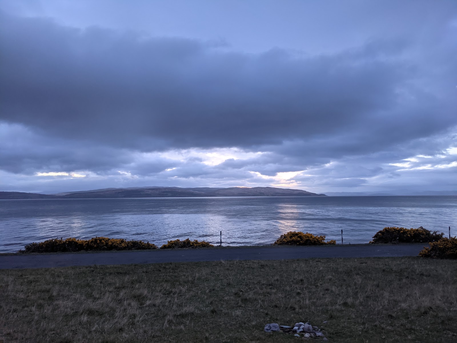 Grey clouds break over hills across a calm sea loch at dusk
