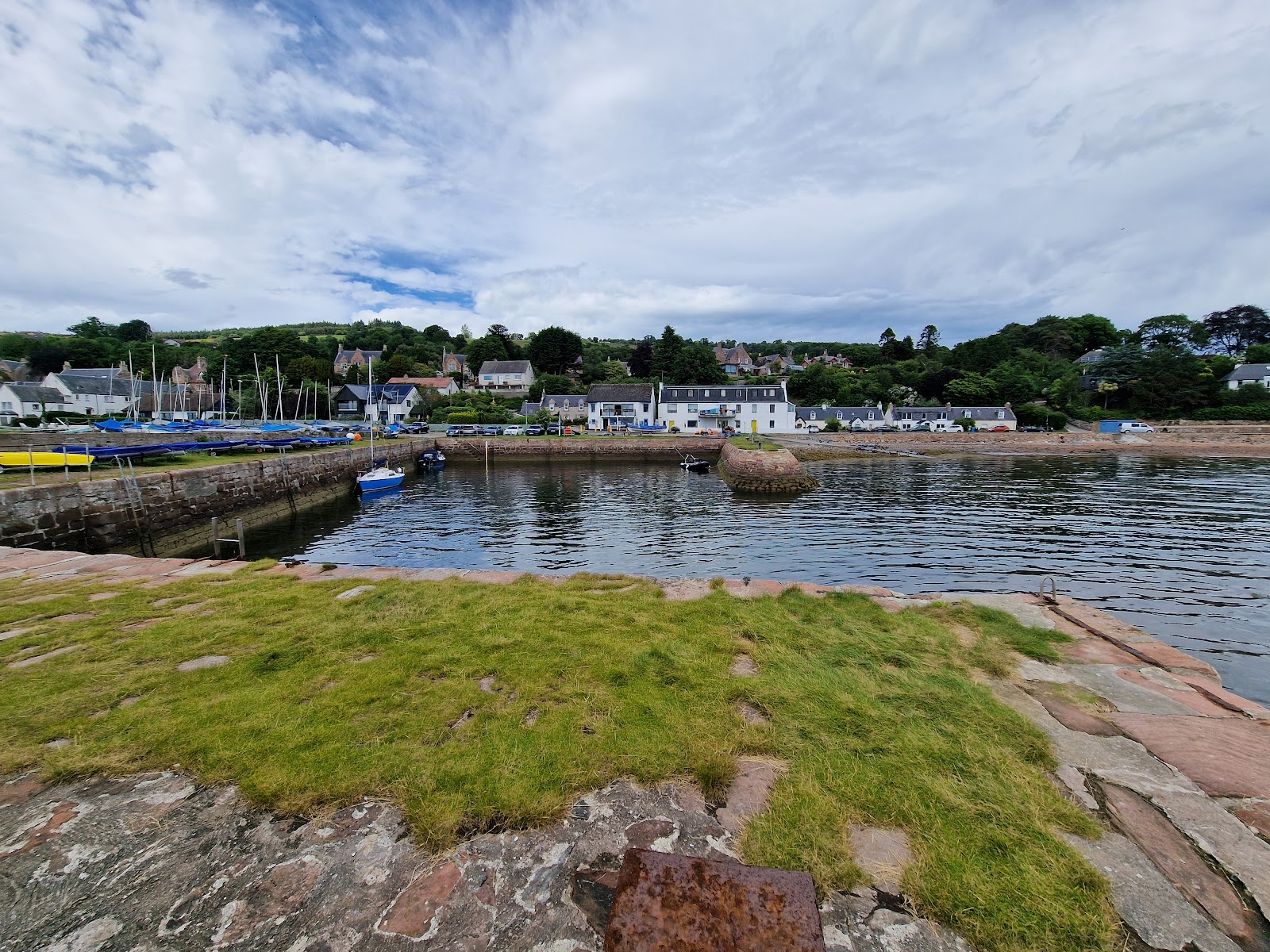 Grassy harbour wall overlooking small boats and white houses along the shore