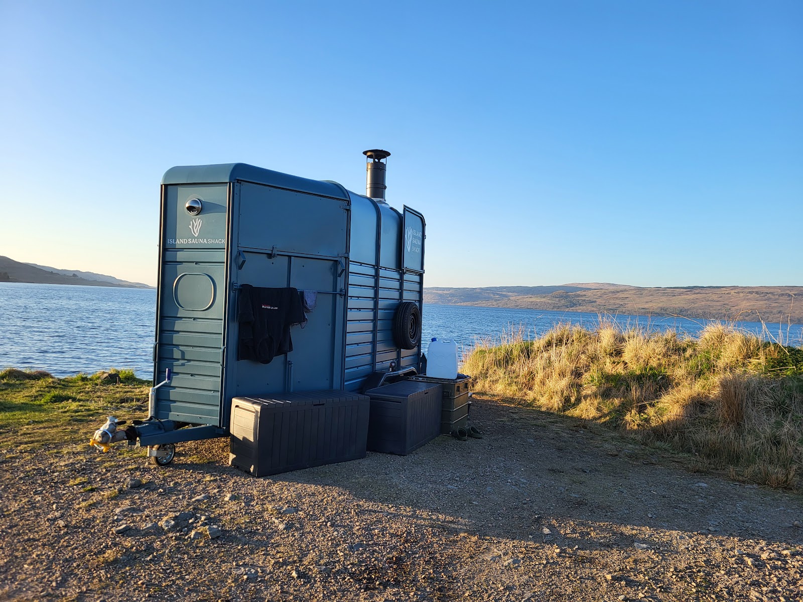 The Island Sauna Shack, a blue converted horsebox with a stove chimney, on a rocky shore by the sea.
