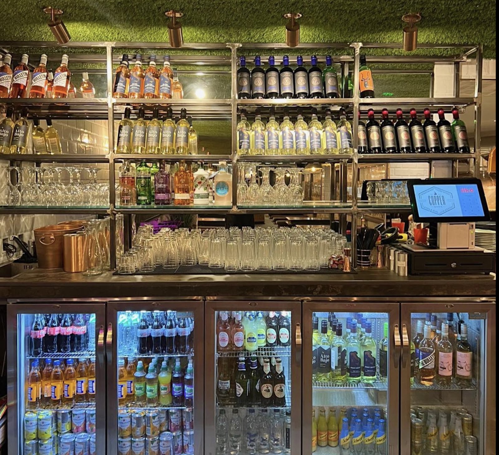 Bar shelves stocked with wine and spirits above illuminated fridges of soft drinks and beer