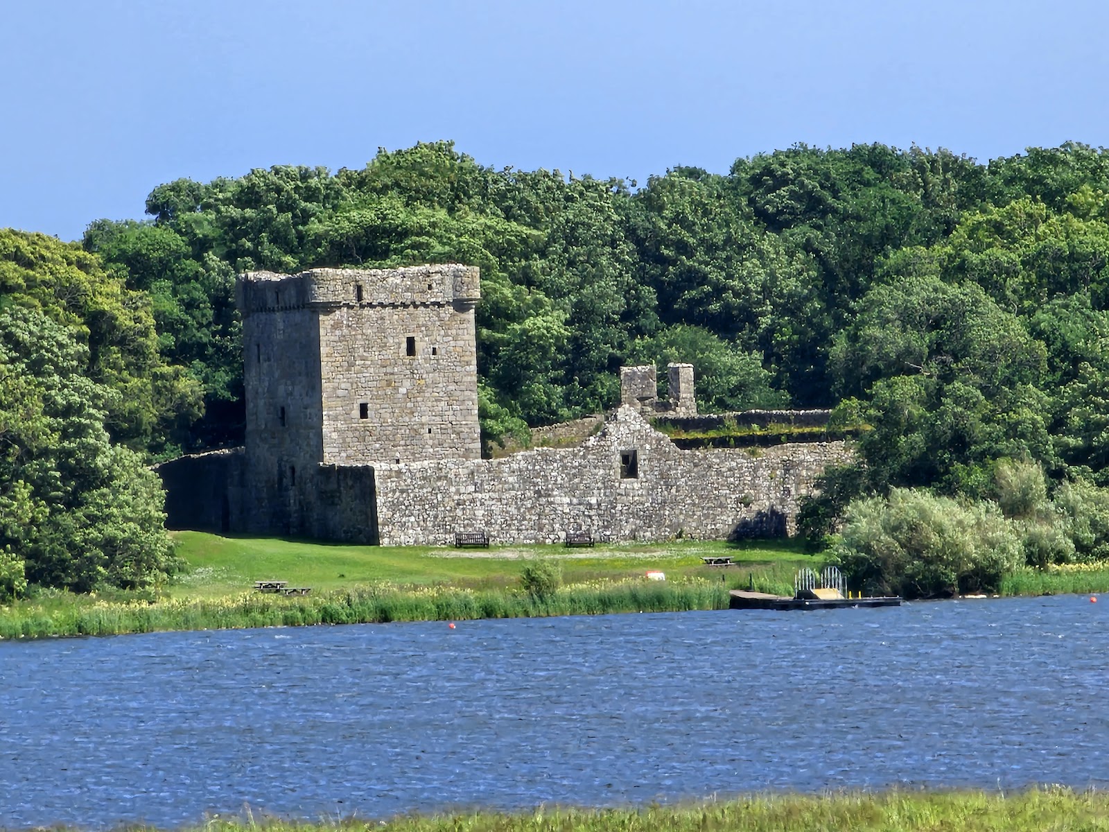 Lochleven Castle, its square stone tower and curtain wall among trees on Castle Island across the blue water of Loch Leven.