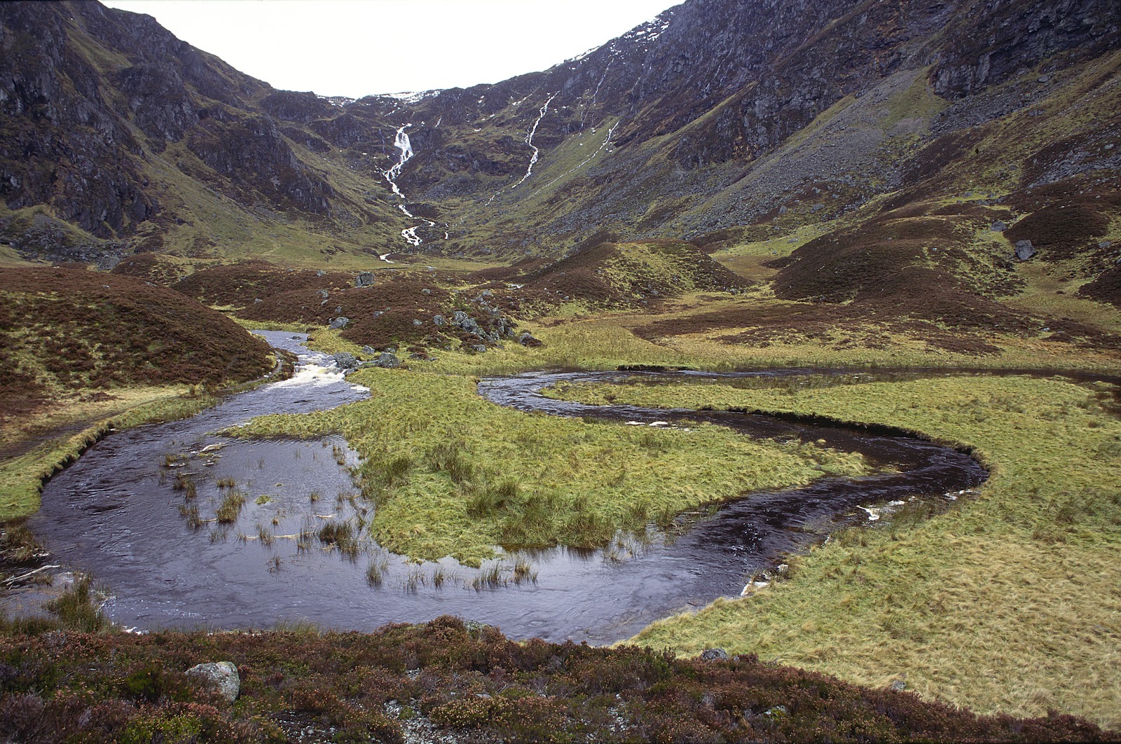 A river meanders through a glen below waterfalls streaking down steep mountainsides