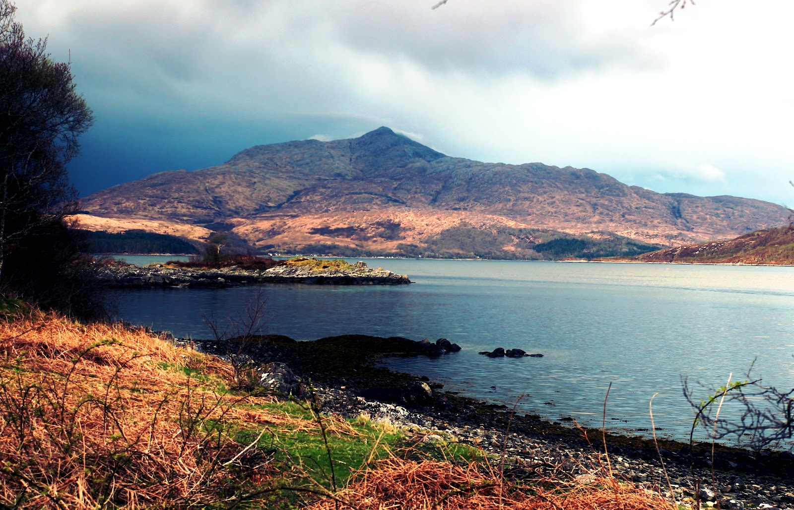 A rocky headland juts into a loch beneath a cloud-covered mountain