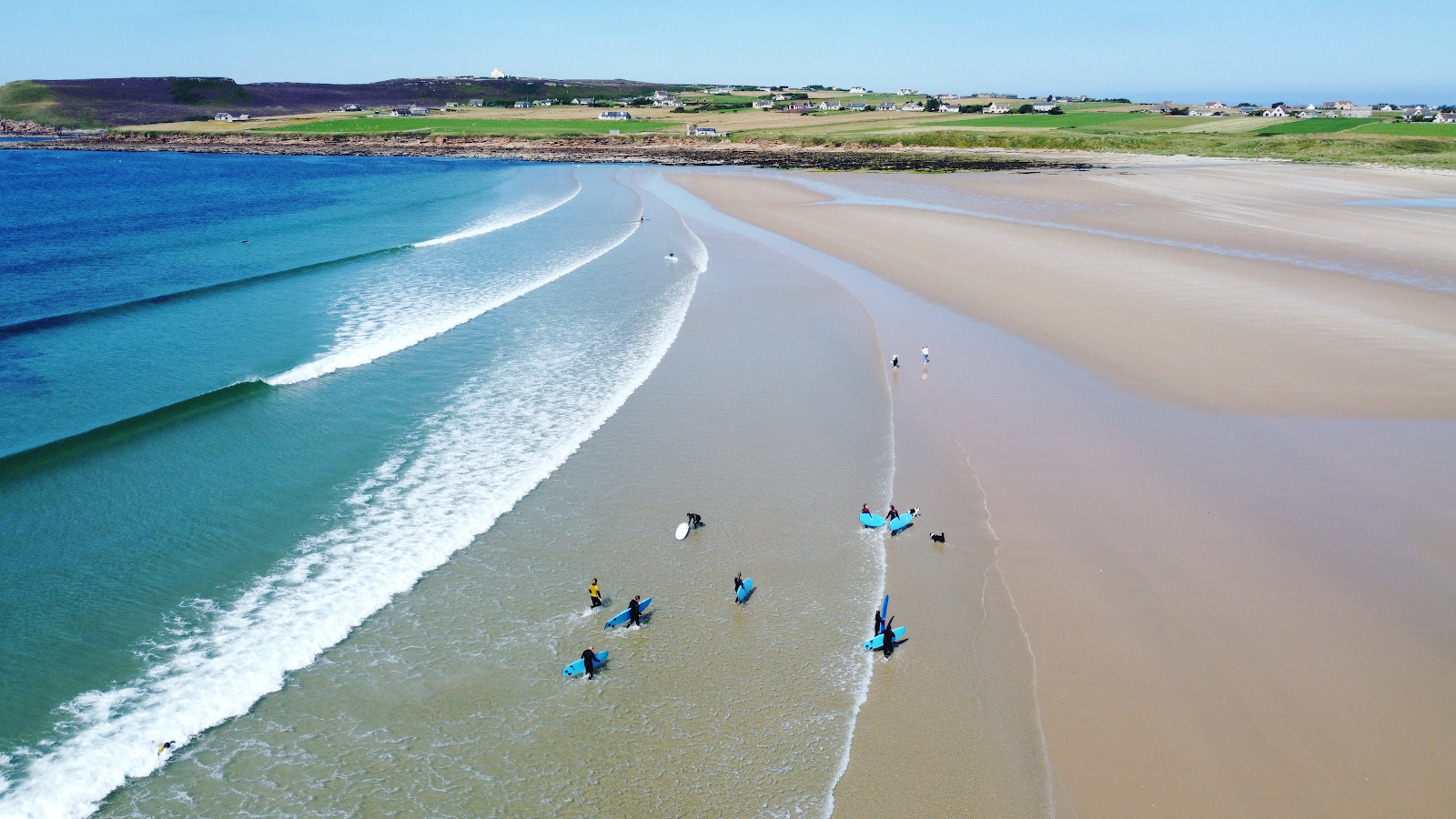 A surf lesson on Dunnet beach seen from the air, learners on boards in the white water along the long sands.