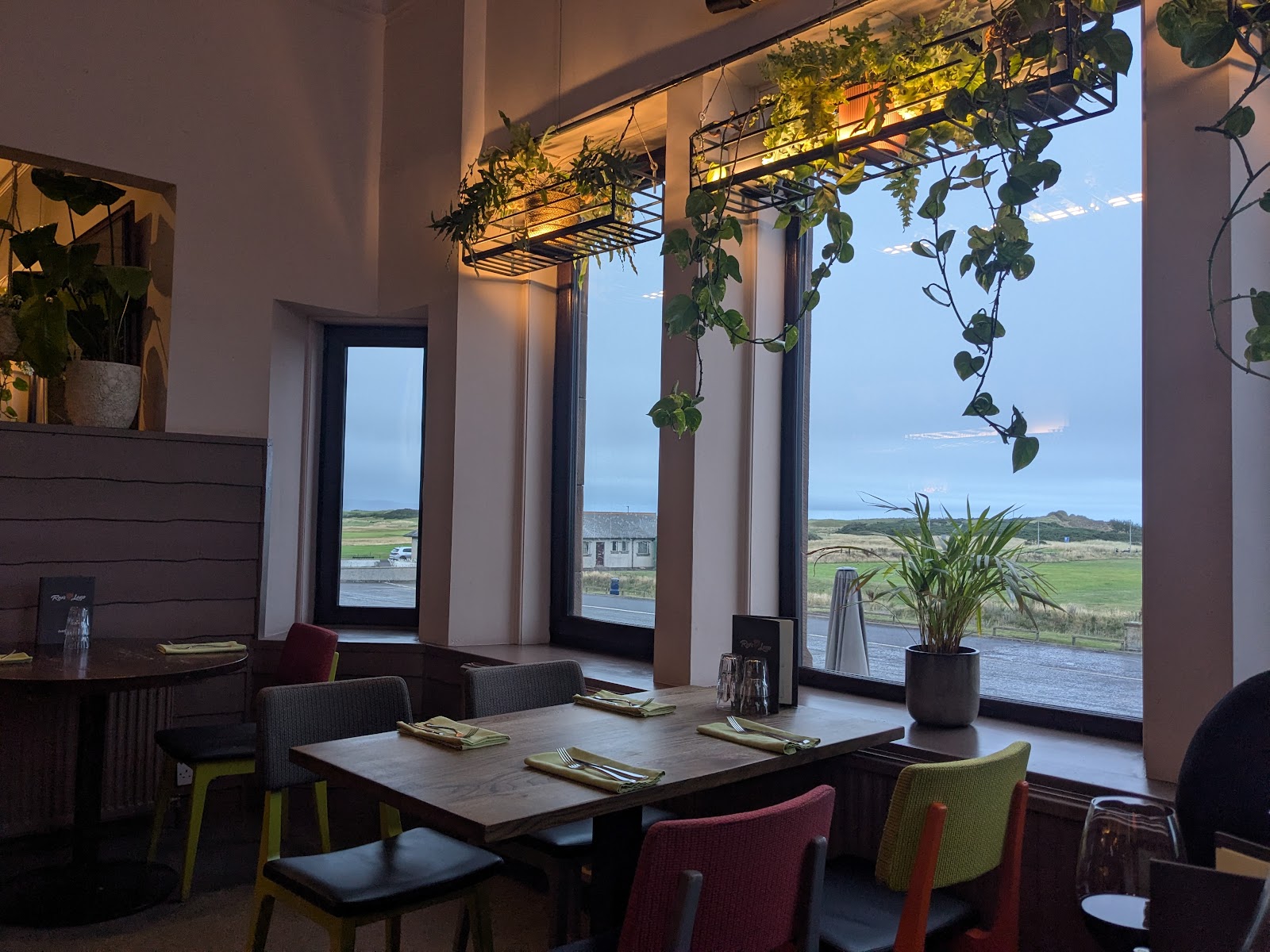 Restaurant tables laid with cutlery beneath hanging plants, overlooking coastal dunes at dusk