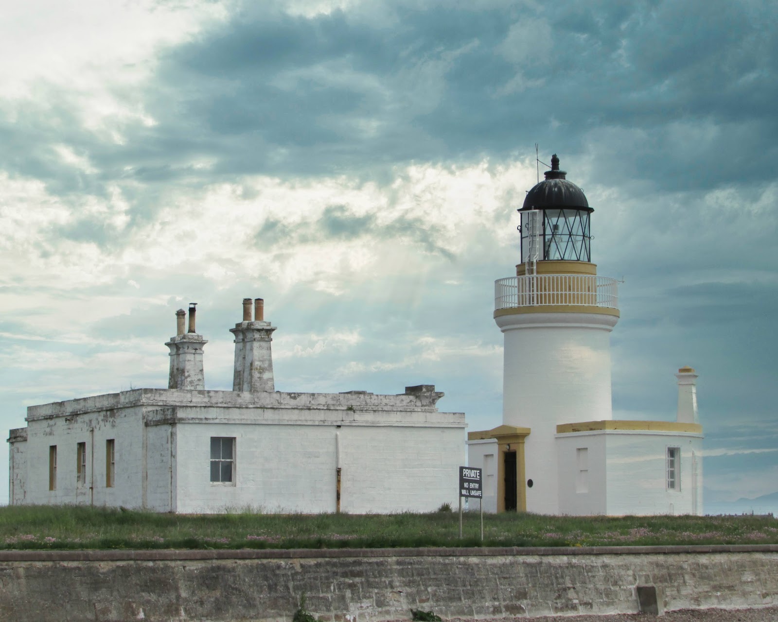 Cromarty lighthouse's white tower and keepers' cottages behind the sea wall under a stormy sky.