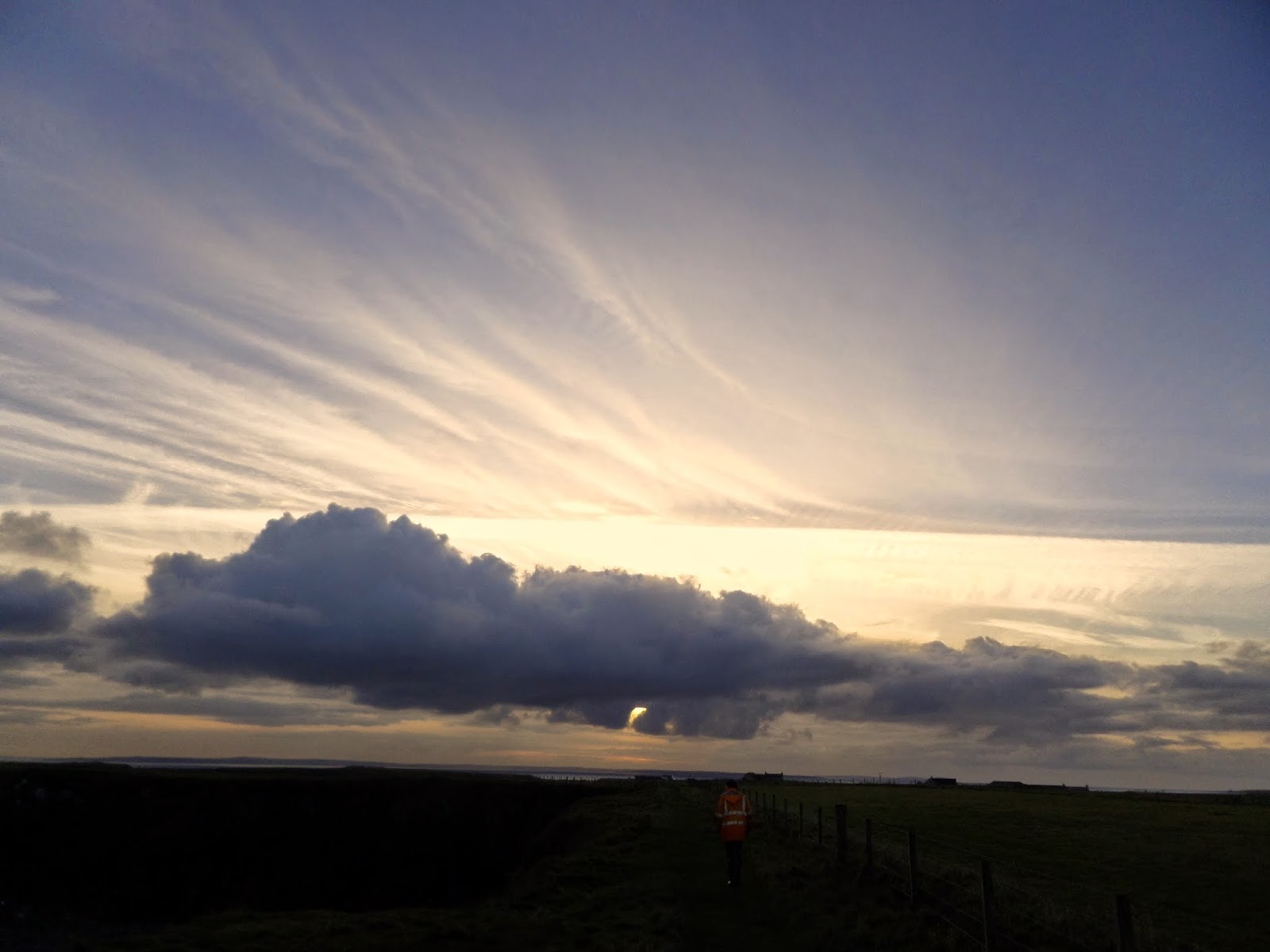 Walker in orange jacket crosses a clifftop field beneath streaked clouds