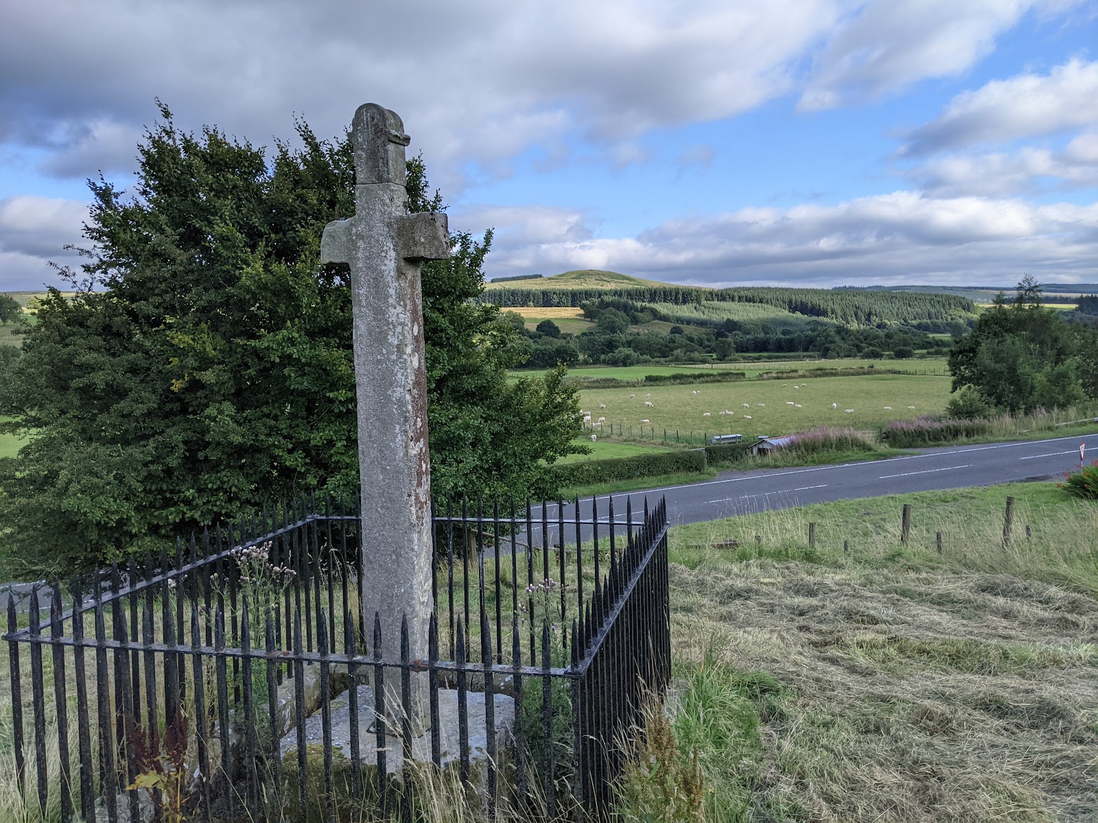 The Milnholm Cross, a tall stone cross inside iron railings in a roadside field, green hills behind.