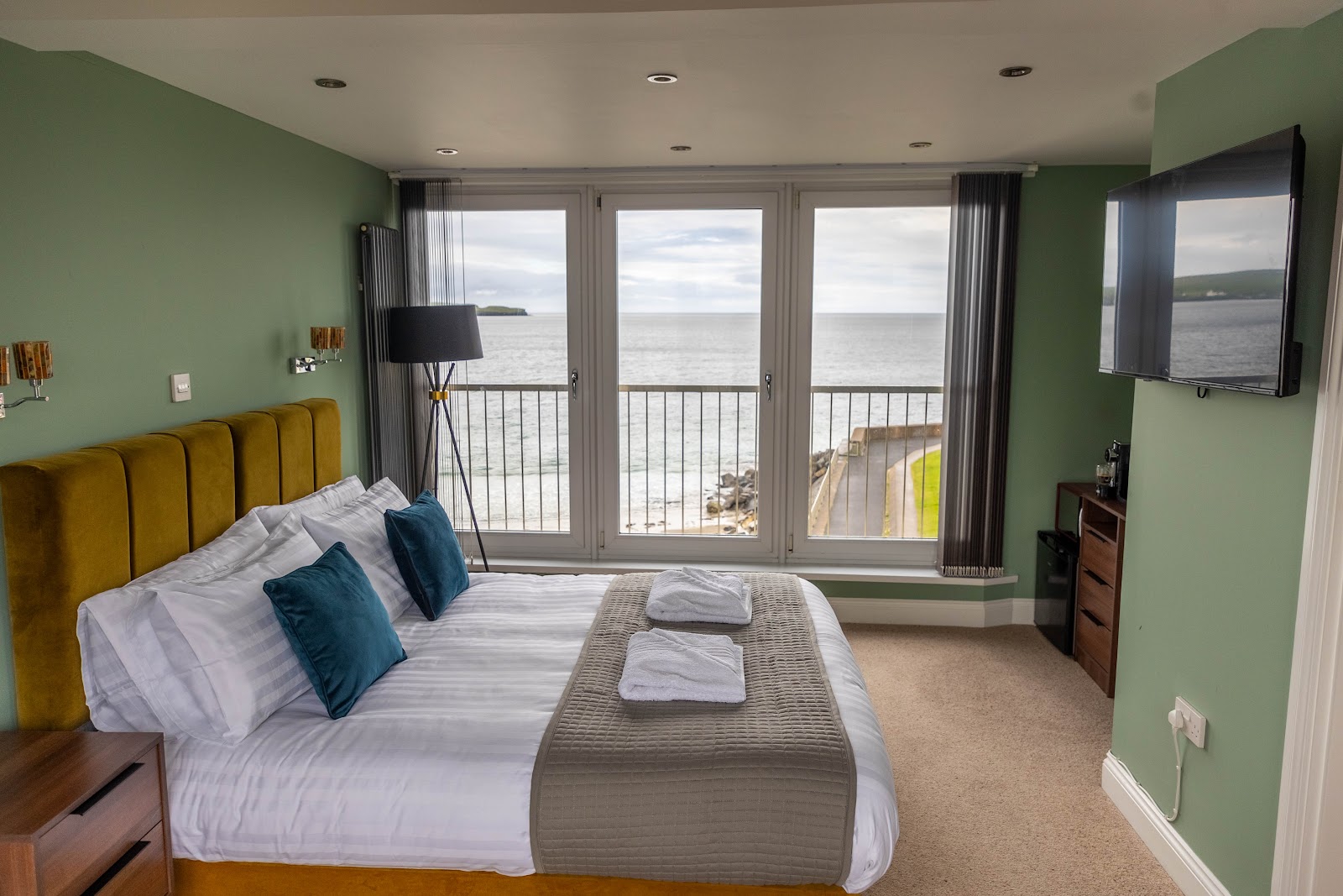 A sea-view room at The View in Thurso, a bed with a mustard headboard facing tall windows over the bay.