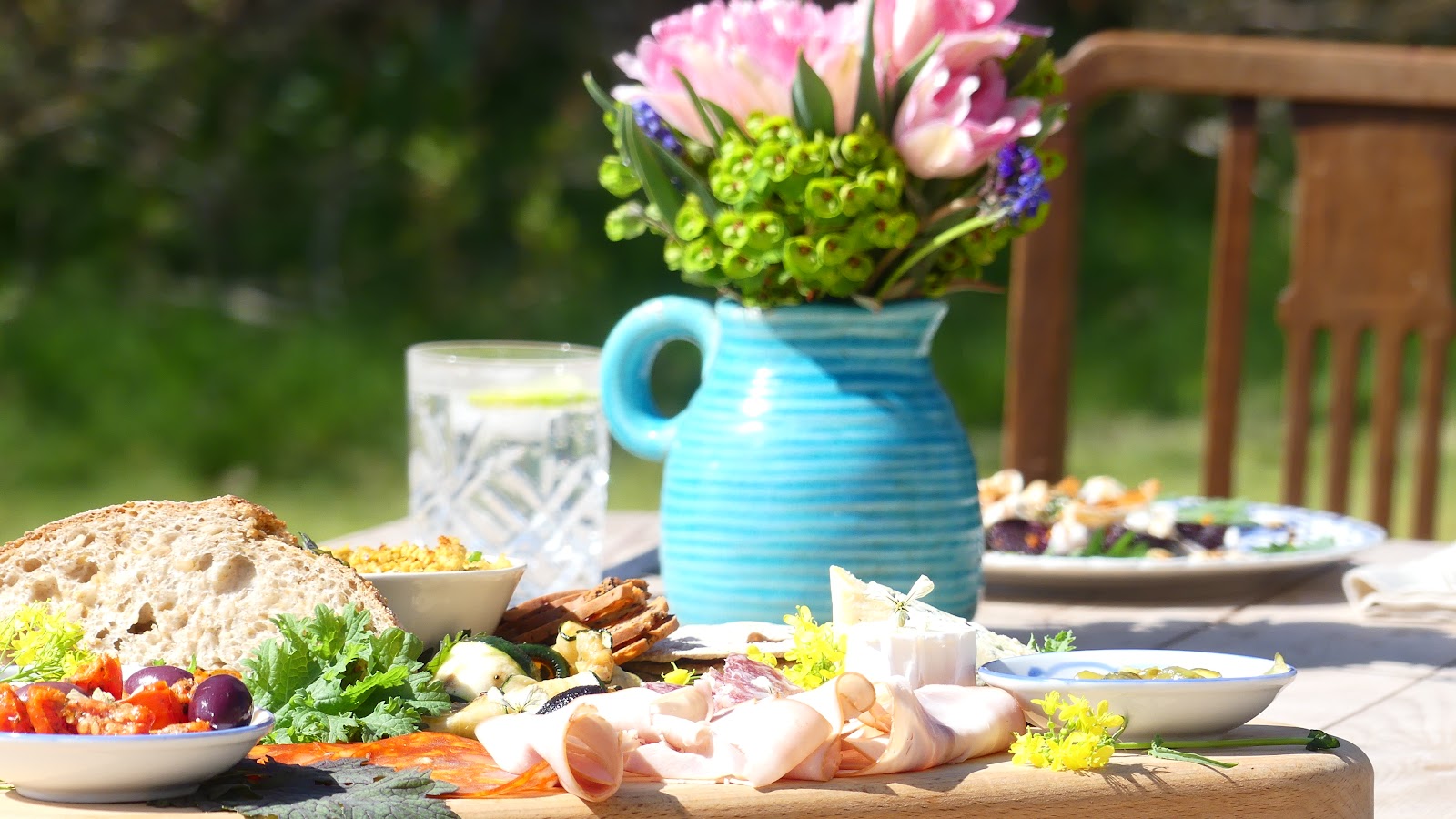 A sharing platter laid on an outdoor table at Wilson's Farm and Kitchen: sourdough, cured meats, salad, cheese and pickles, beside a blue jug of pink and green flowers.