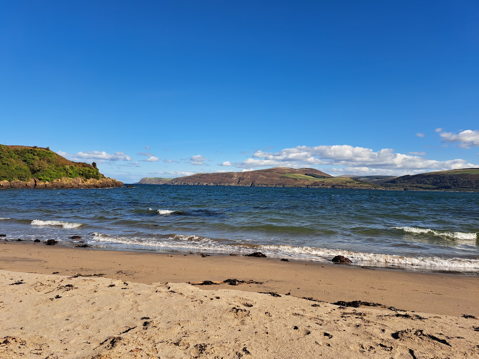 The sandy beach at Lady Bay with small waves breaking, Loch Ryan beyond and the hills of its far shore under a deep blue sky, a wooded headland on the left.