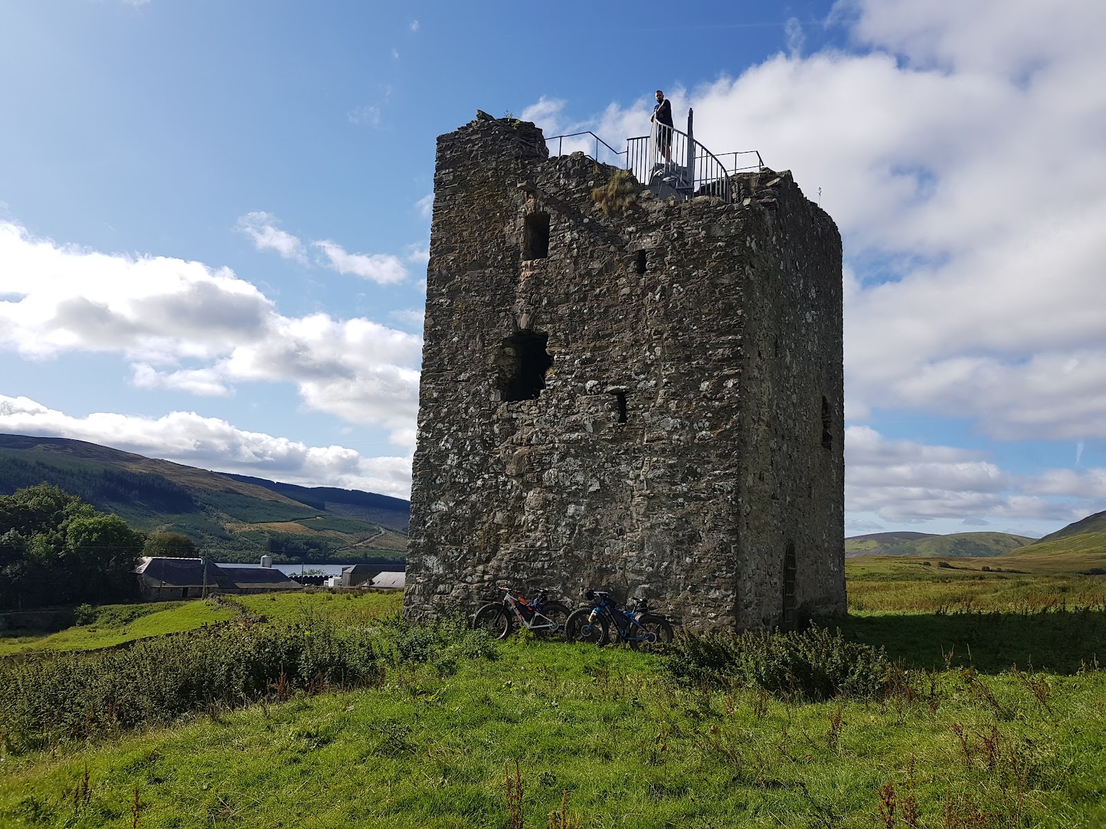 Dryhope Tower in sunshine, a roofless stone tower house with a railed platform on top, bicycles at its foot and St Mary's Loch and Dryhope farm beyond.