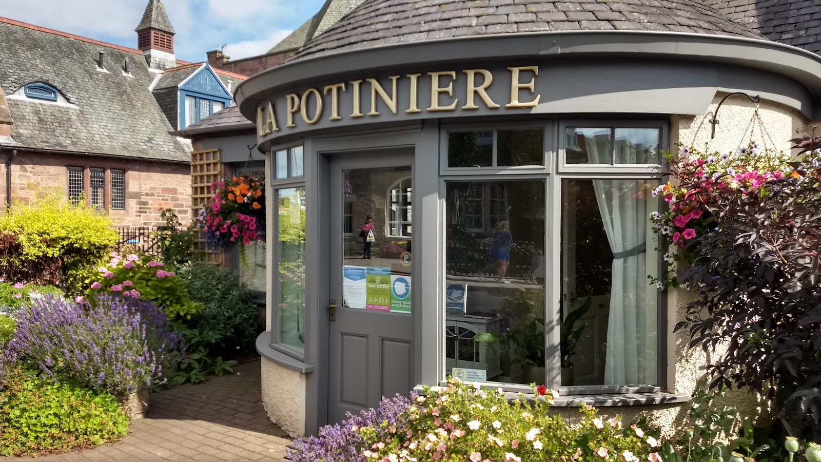 The front of La Potinière on Main Street in Gullane, a curved grey-painted shopfront with its name in gold letters above the door, lavender and summer flowers in bloom outside.