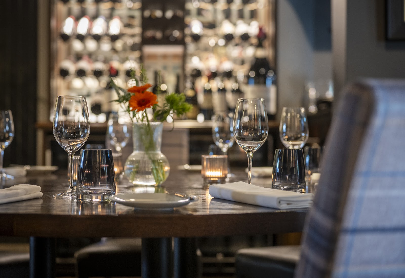 A table laid at Amuse with wine glasses, white napkins, a candle and a vase of orange flowers, in front of a wall of wine bottles.