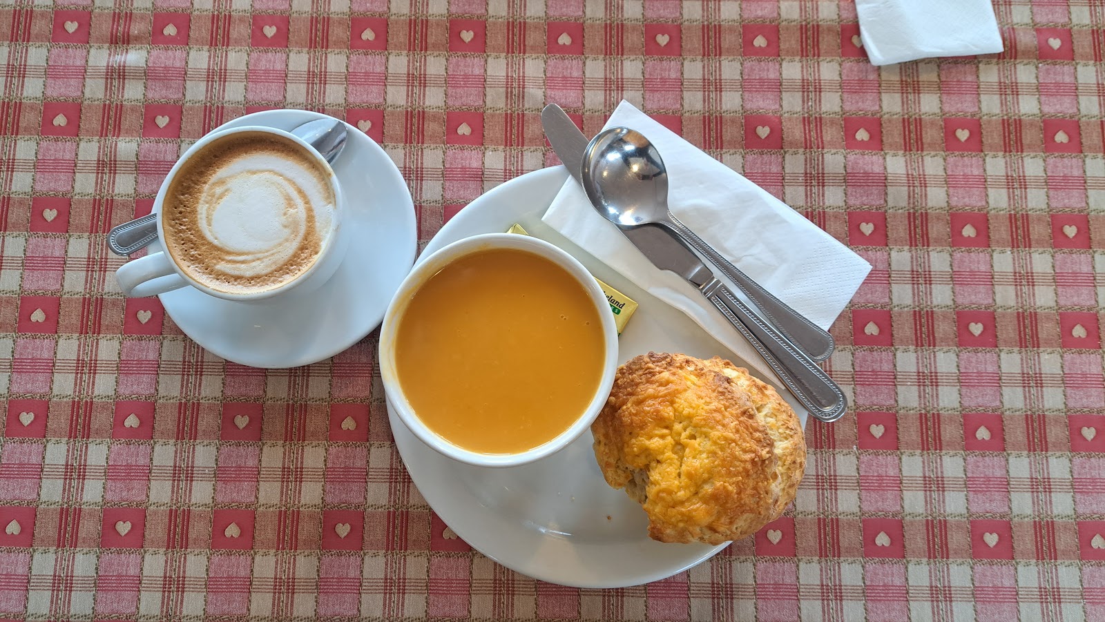 A bowl of orange soup with a cheese scone and a cappuccino on a red-checked tablecloth at The Joinery in Meigle.