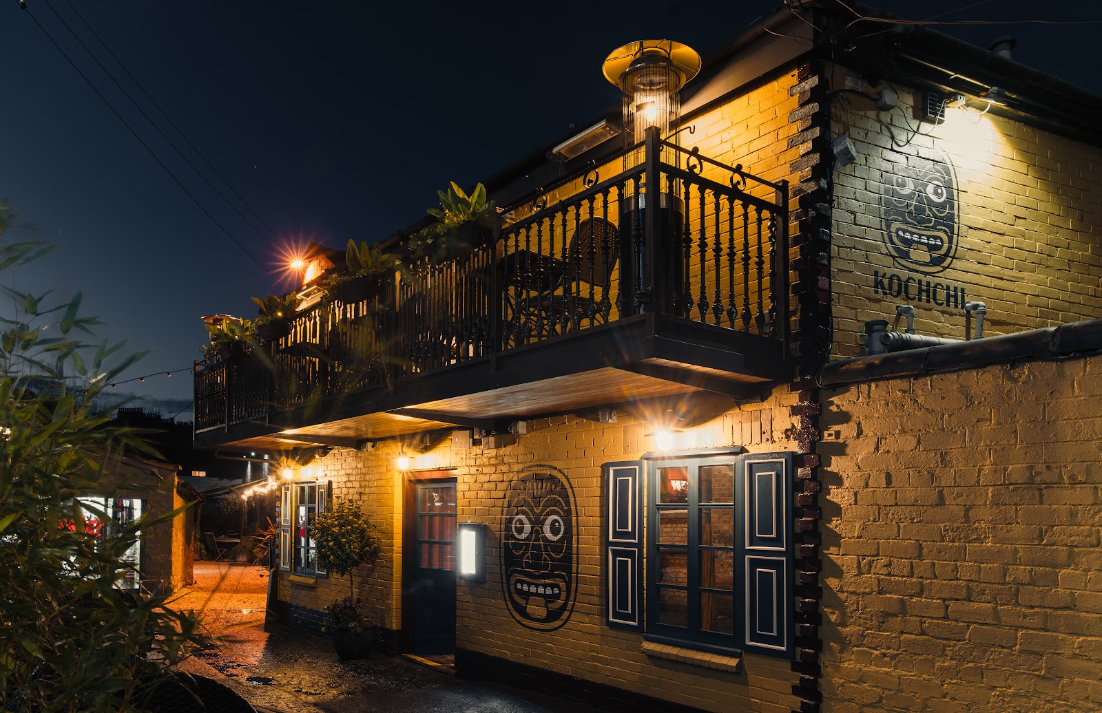Kochchi's yellow brick building on Ruthven Lane at night, with its balcony lit above and its masks painted on the walls.