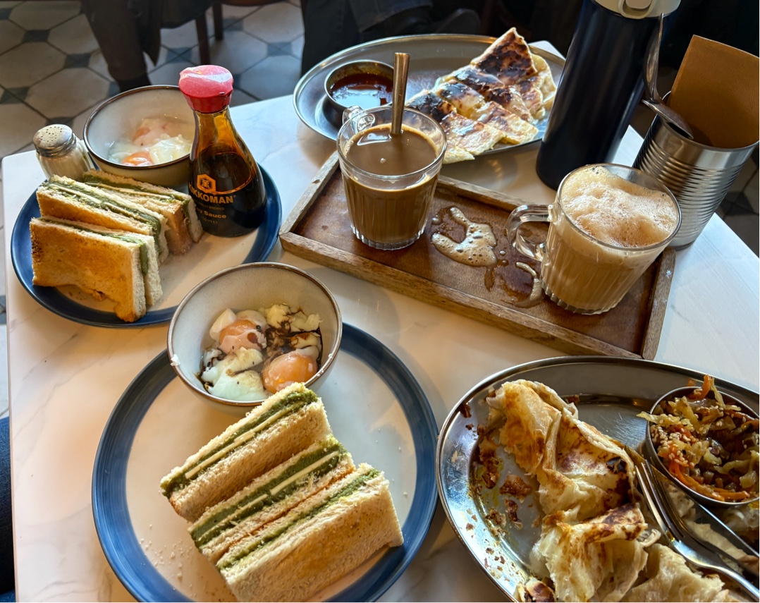 Kaya toast, soft-boiled eggs with soy sauce, roti with curry and two glasses of kopi on a marble table at Singapore Coffee House.