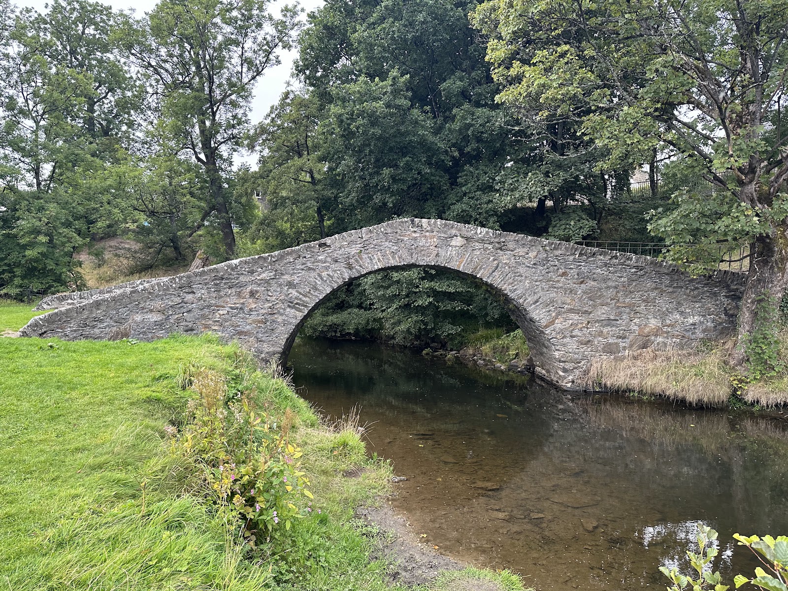 The Auld Brig's single stone arch over the slow, clear River Isla in Keith, between grassy banks and trees in leaf.