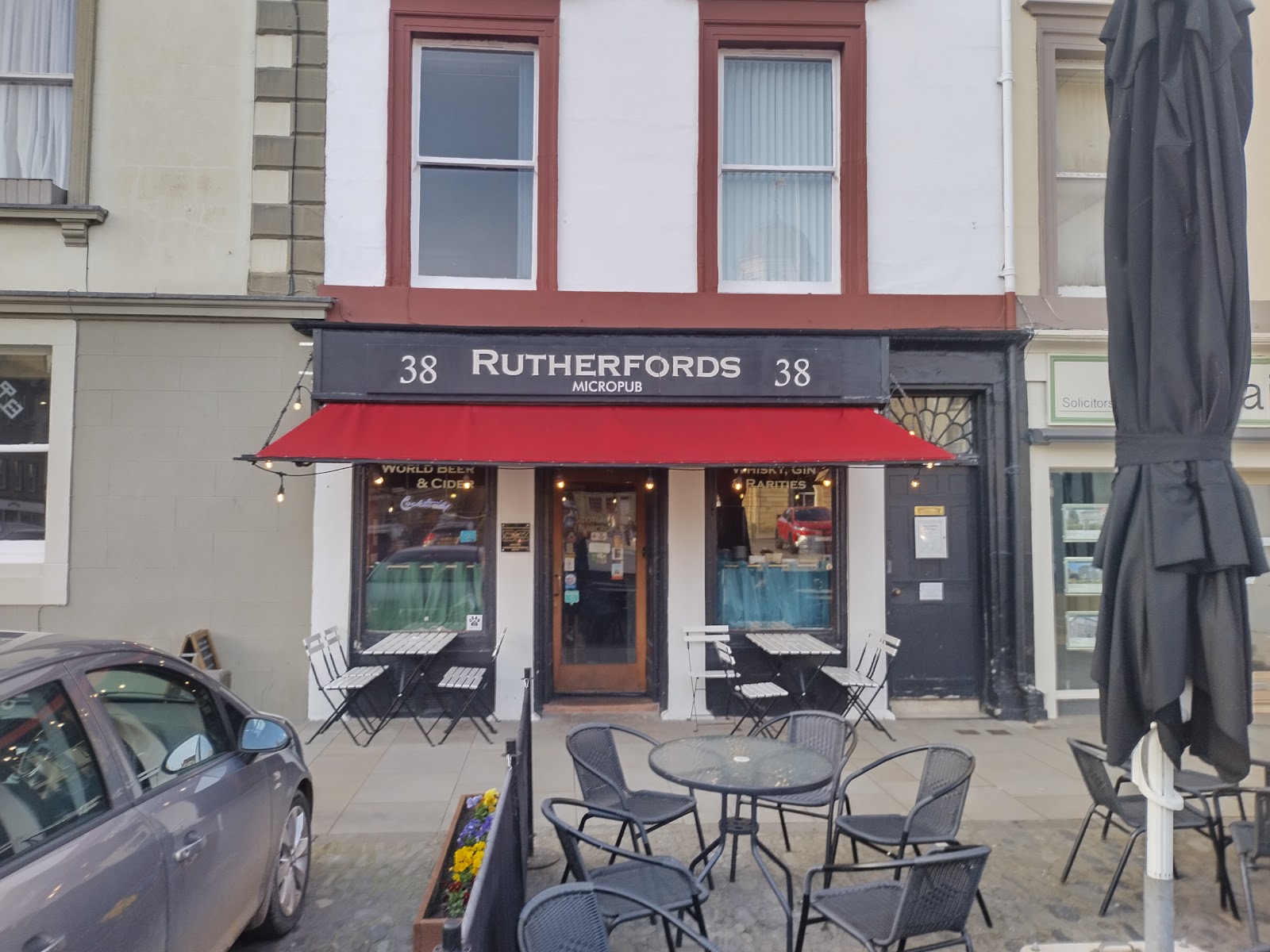 The shopfront of Rutherfords Micropub at 38 The Square in Kelso, with its black sign, red awning and tables and chairs on the pavement.