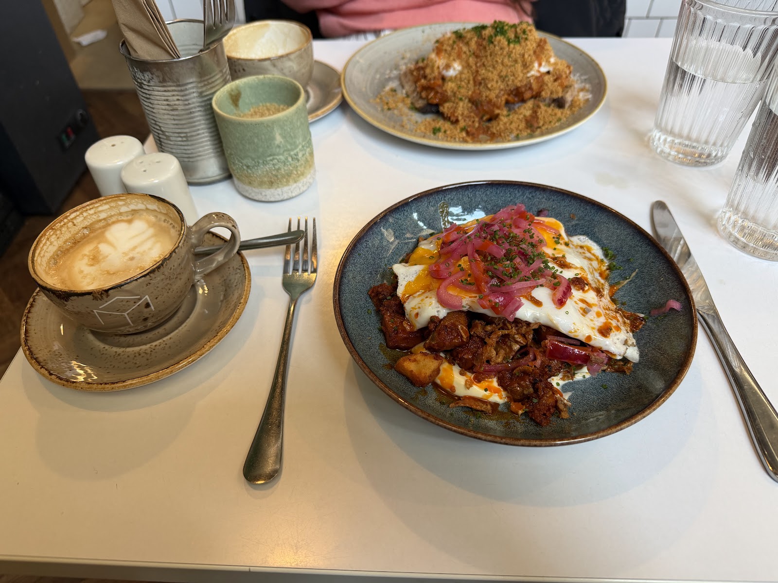 Brunch on a white table at Mesa on Duke Street: fried eggs over a spiced hash with pickled red onion in a blue bowl, a cappuccino beside it and a second plate behind.