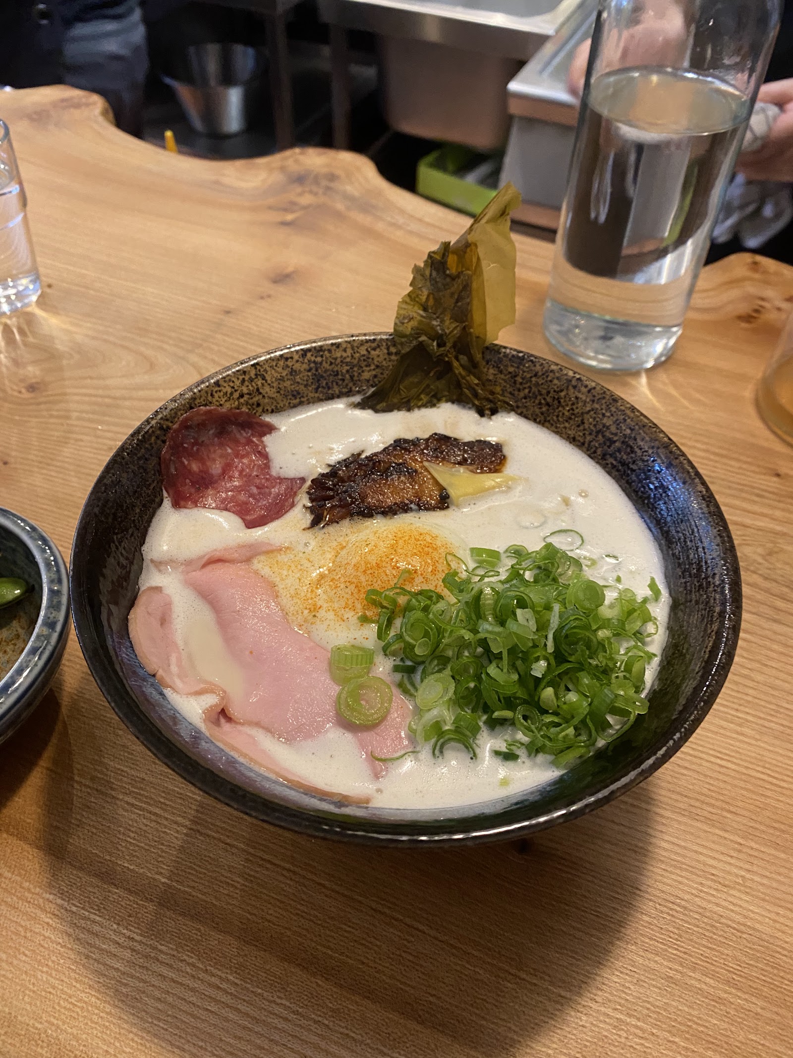 A bowl of tonkotsu ramen at Gulp Ramen, with slices of pork belly, duck, an onsen egg and spring onion in a creamy broth, on a wooden counter.