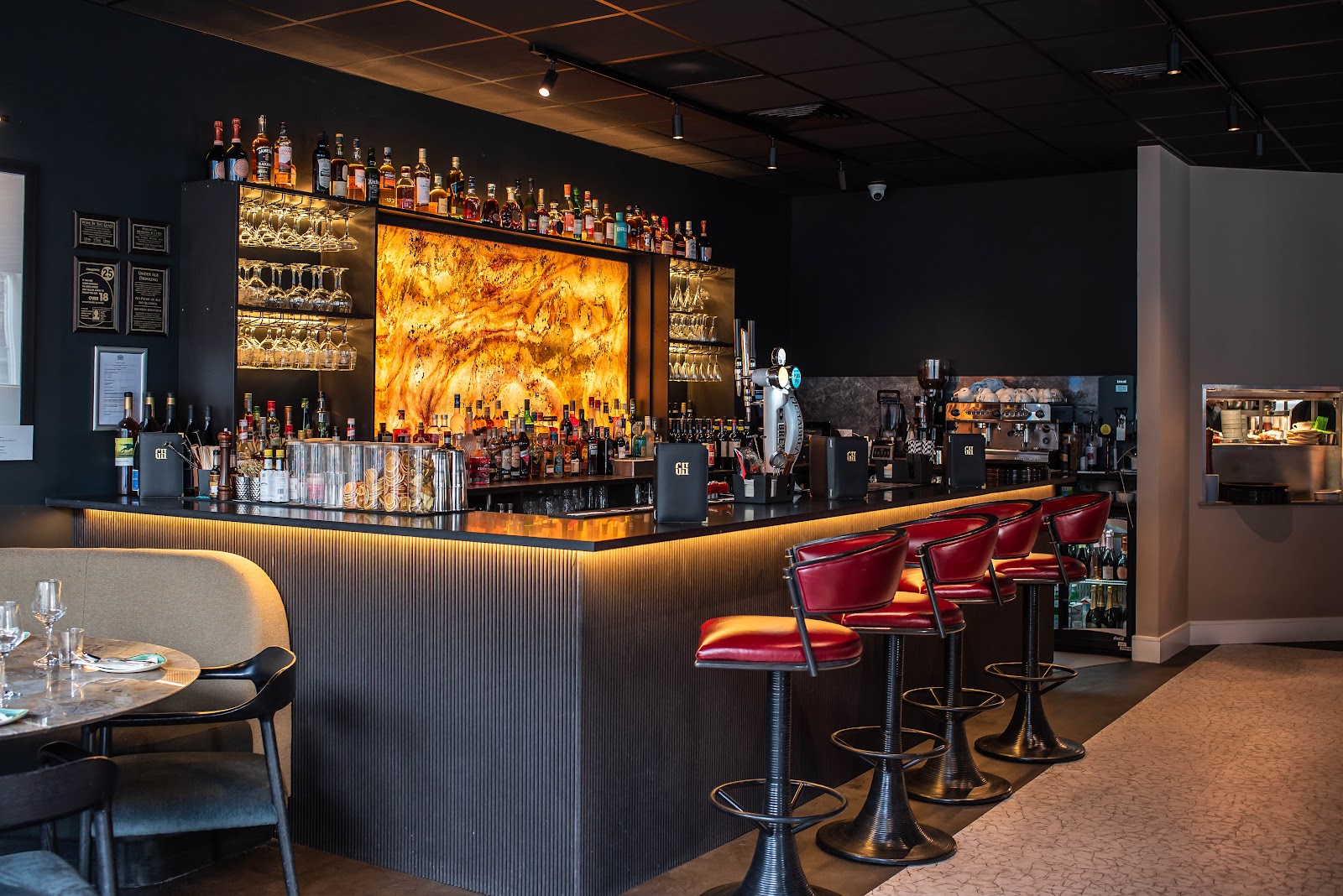 The bar at Glenhouse, lit in amber under a wall of bottles, with red leather stools and a round marble table.