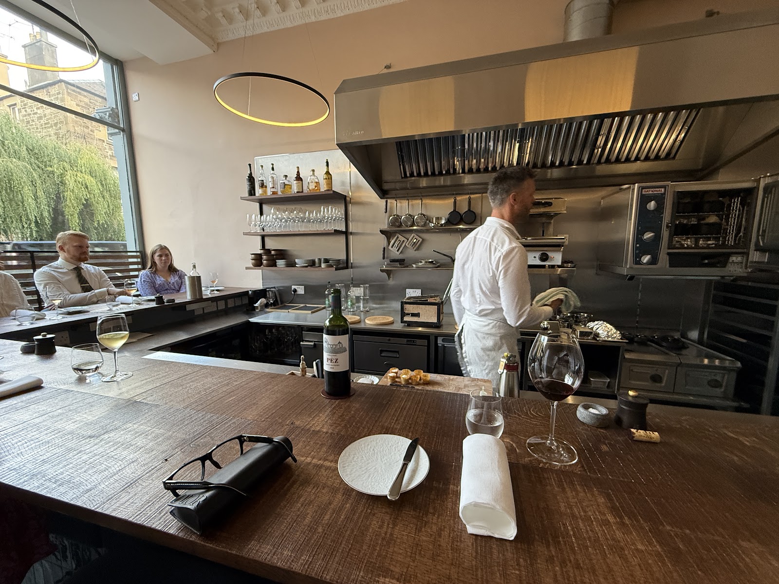 The chef at work in the open kitchen at Argile, seen across the long wooden counter where guests sit, with a wine glass and a folded napkin laid in front.
