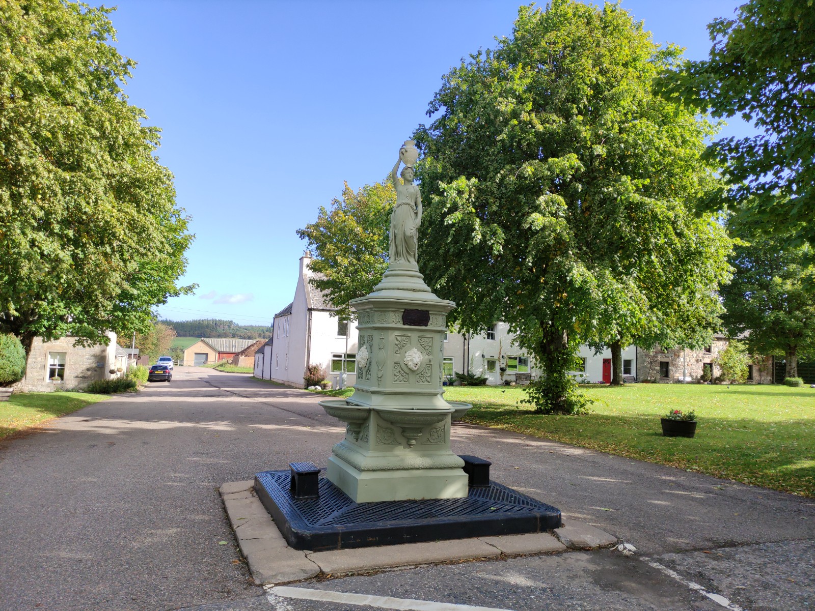 The Square in Tomintoul, a green drinking fountain with a figure on top between trees and stone houses.