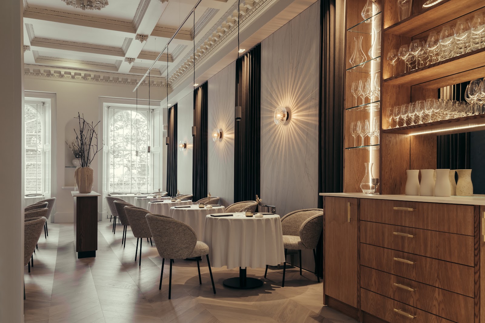 The dining room at LYLA on Royal Terrace, round tables laid with white cloths under a tall arched window, with lamps glowing on the pale walls and a glass-shelved bar to one side.