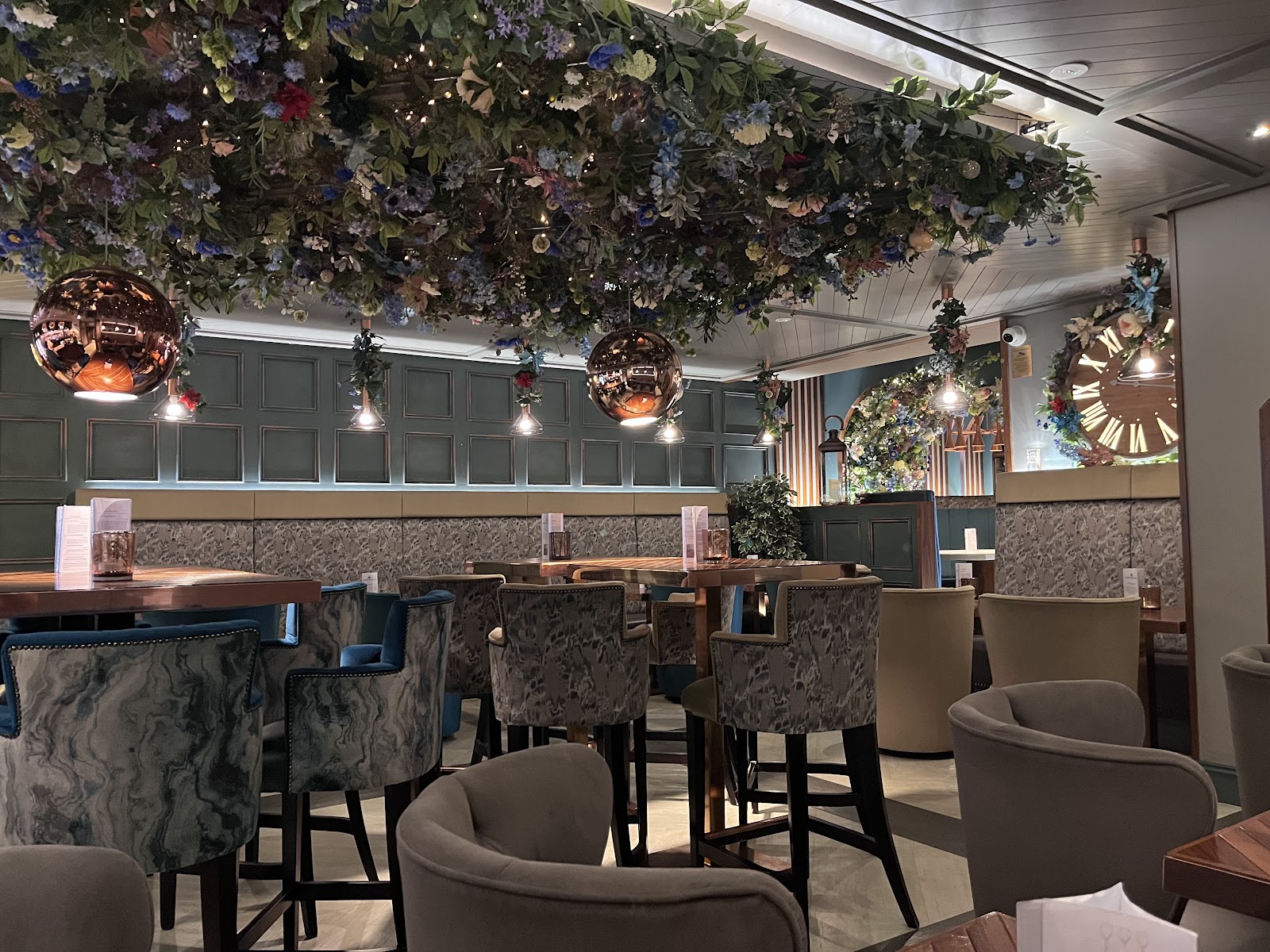 The bar room at Grape & Grain, under a ceiling hung with flowers and copper lamps, with high stools, velvet tub chairs and a large clock on the far wall.