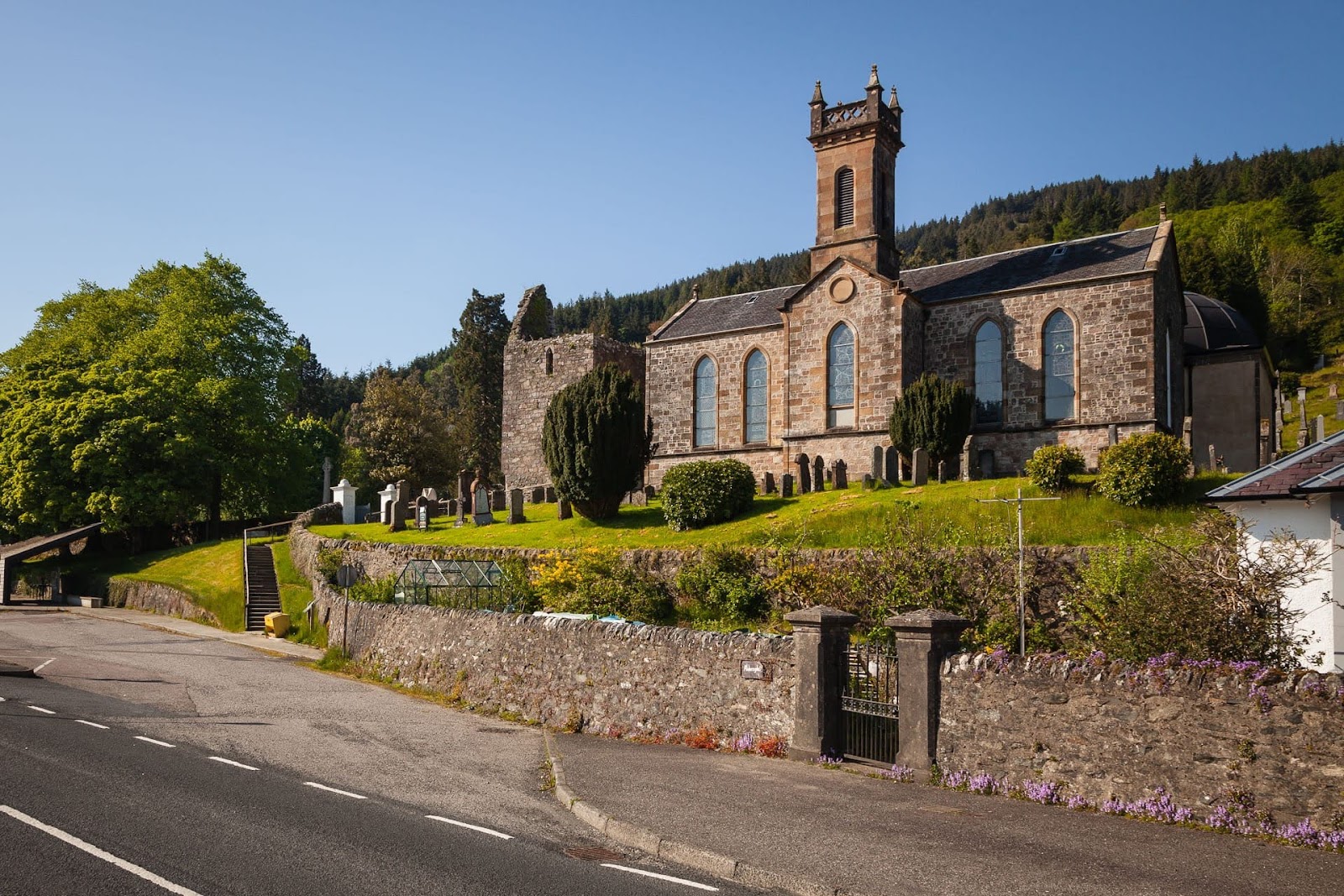 St Munn's Church at Kilmun in sunshine, its square tower and tall windows above the sloping churchyard and its stone wall, the ruined medieval tower beside it and the wooded hill behind.