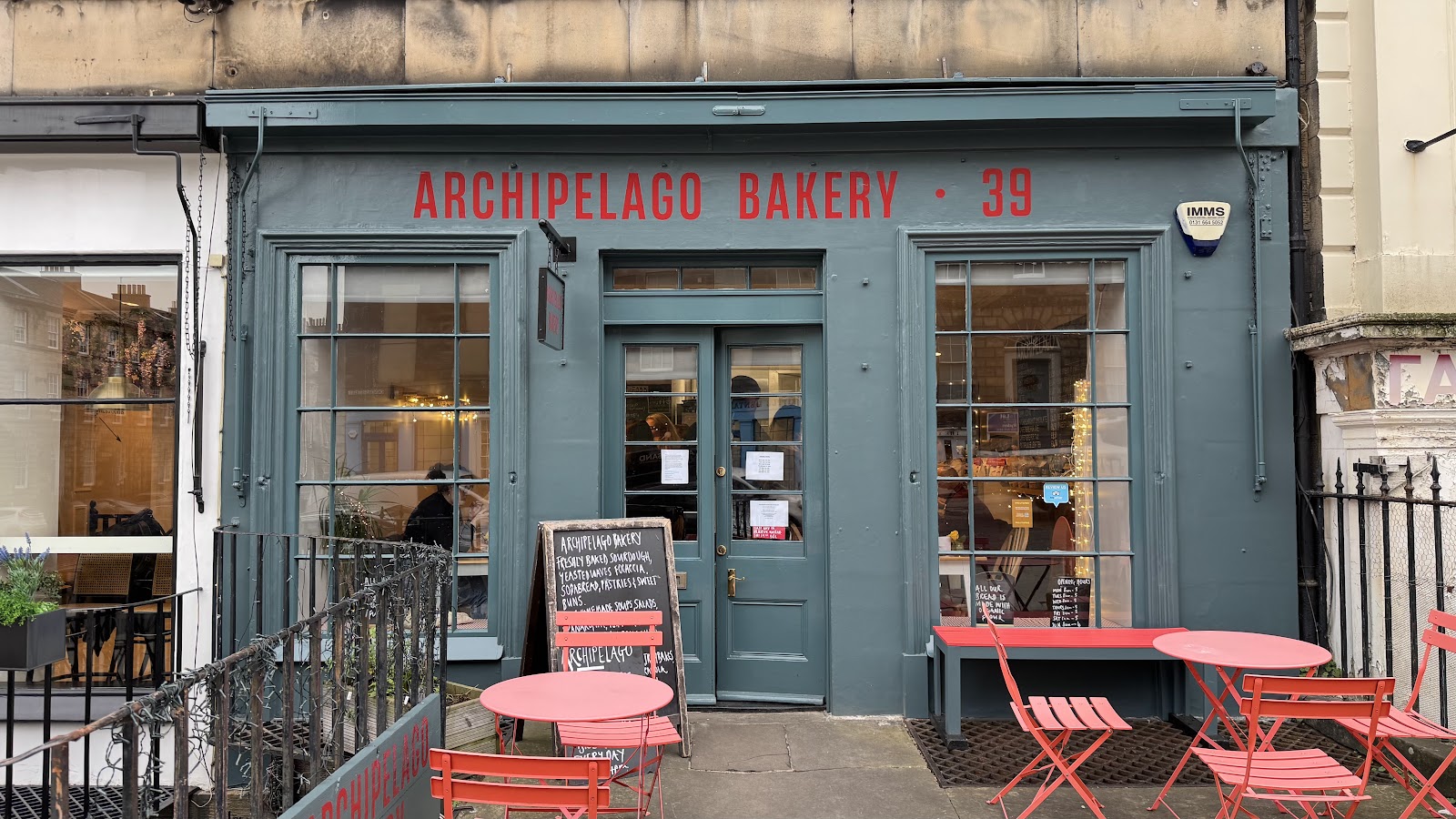 The grey-green front of Archipelago Bakery at 39 Dundas Street, with its name in red above the door, a chalkboard outside and pink tables and chairs on the pavement.