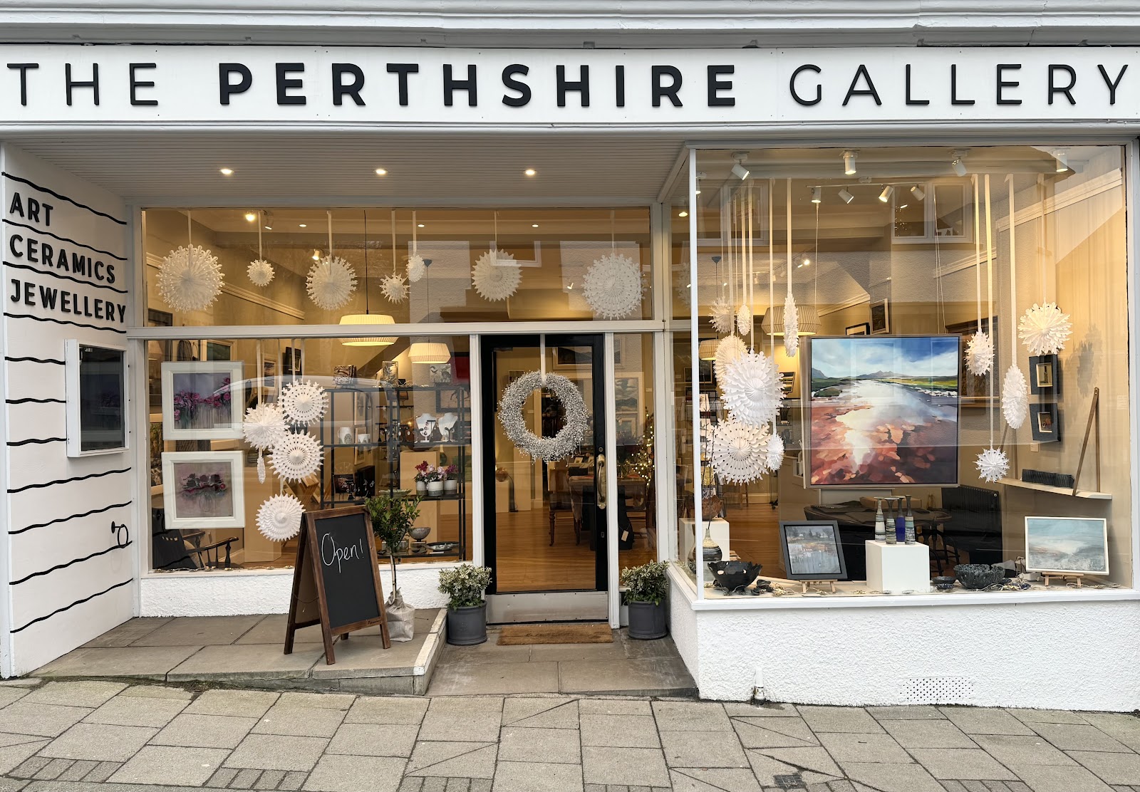The white shopfront of the Perthshire Gallery in Pitlochry, its windows full of paintings and hanging lights.