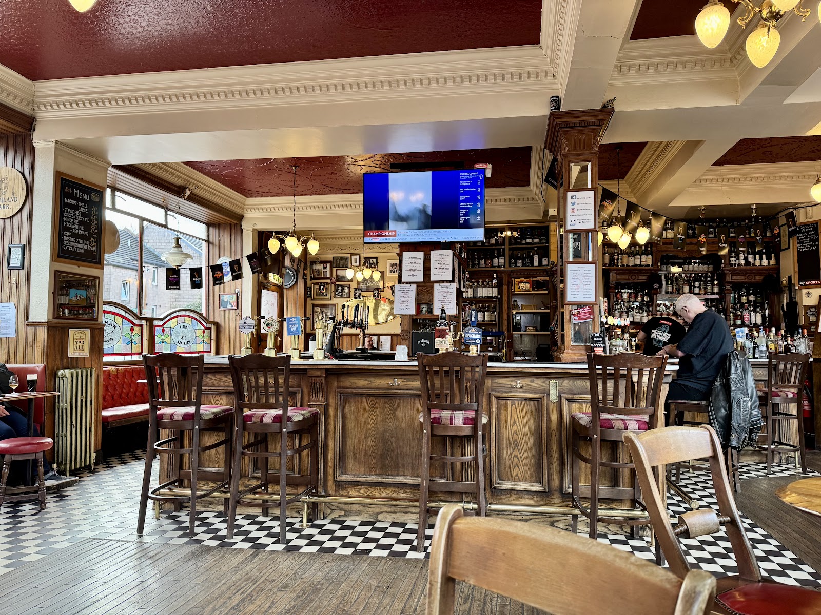The bar of the Athletic Arms in Edinburgh, with tall stools at a wooden counter, a black and white tiled floor and shelves of whisky behind.