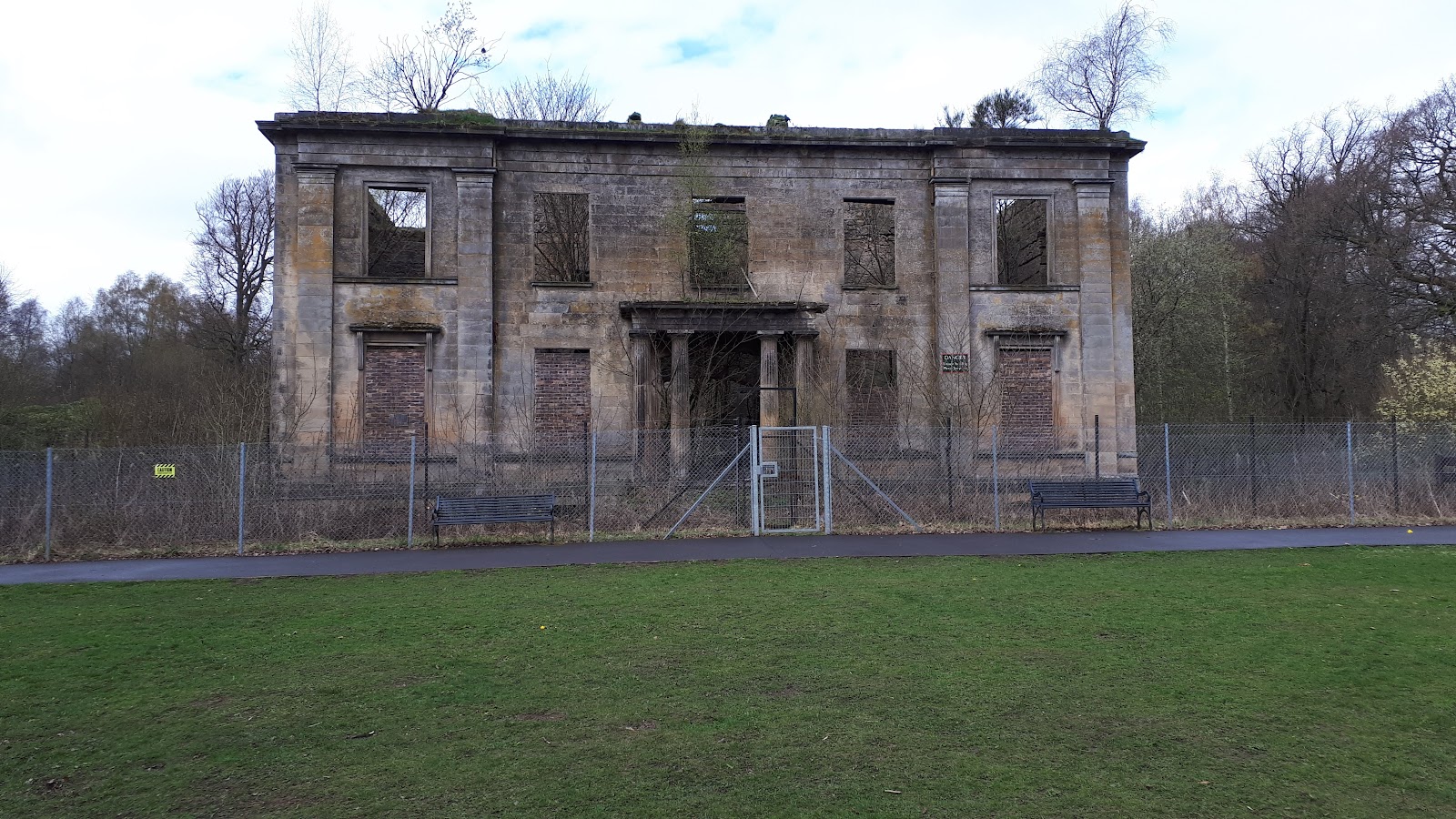 The roofless front of Plean House, its windows empty and its doorway pillared, behind a wire fence, with benches and a lawn in front.