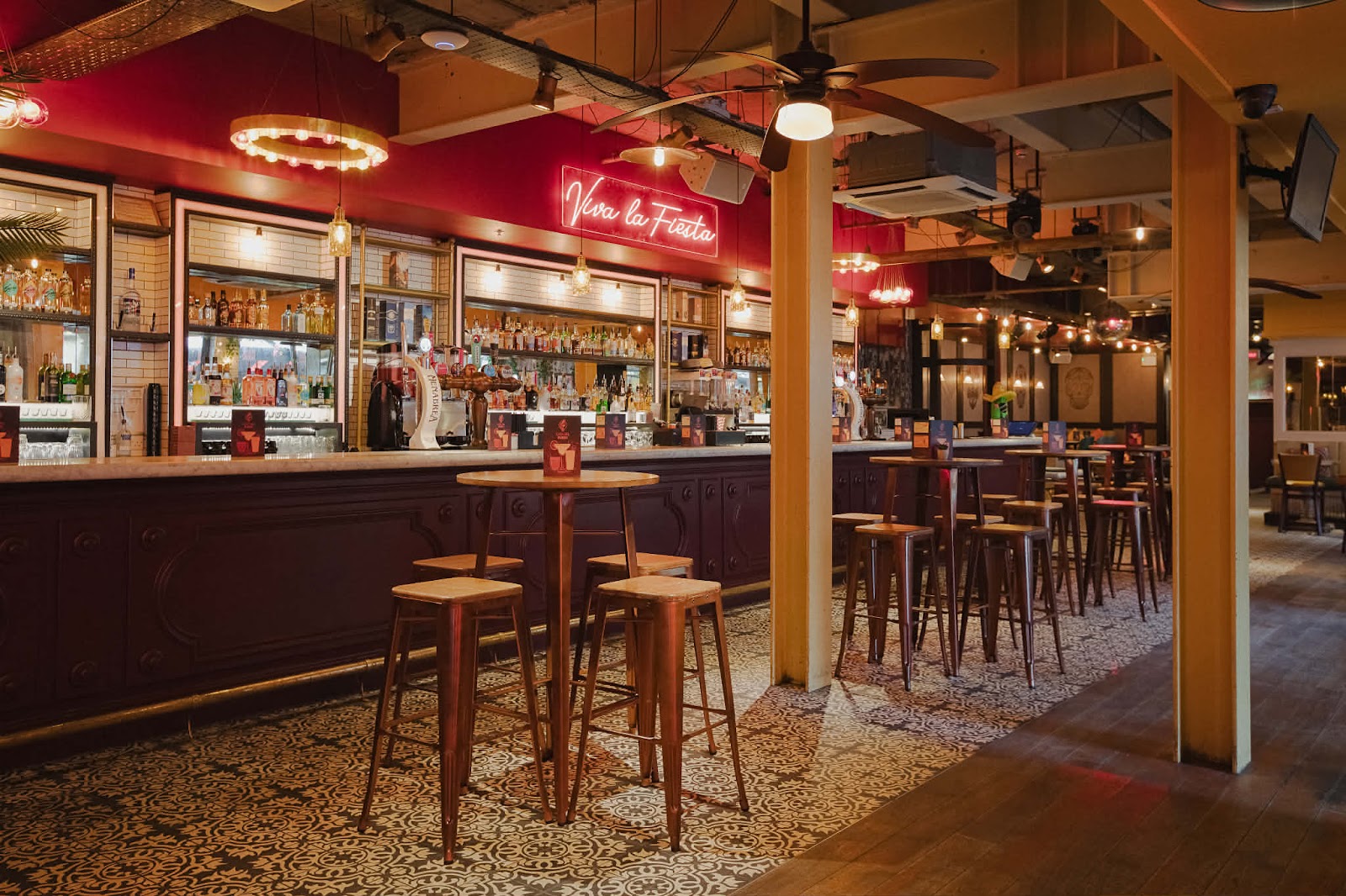 The long bar at Casa del Fuego under a red wall and a neon sign reading Viva la Fiesta, with high stools and tables on a patterned tiled floor.
