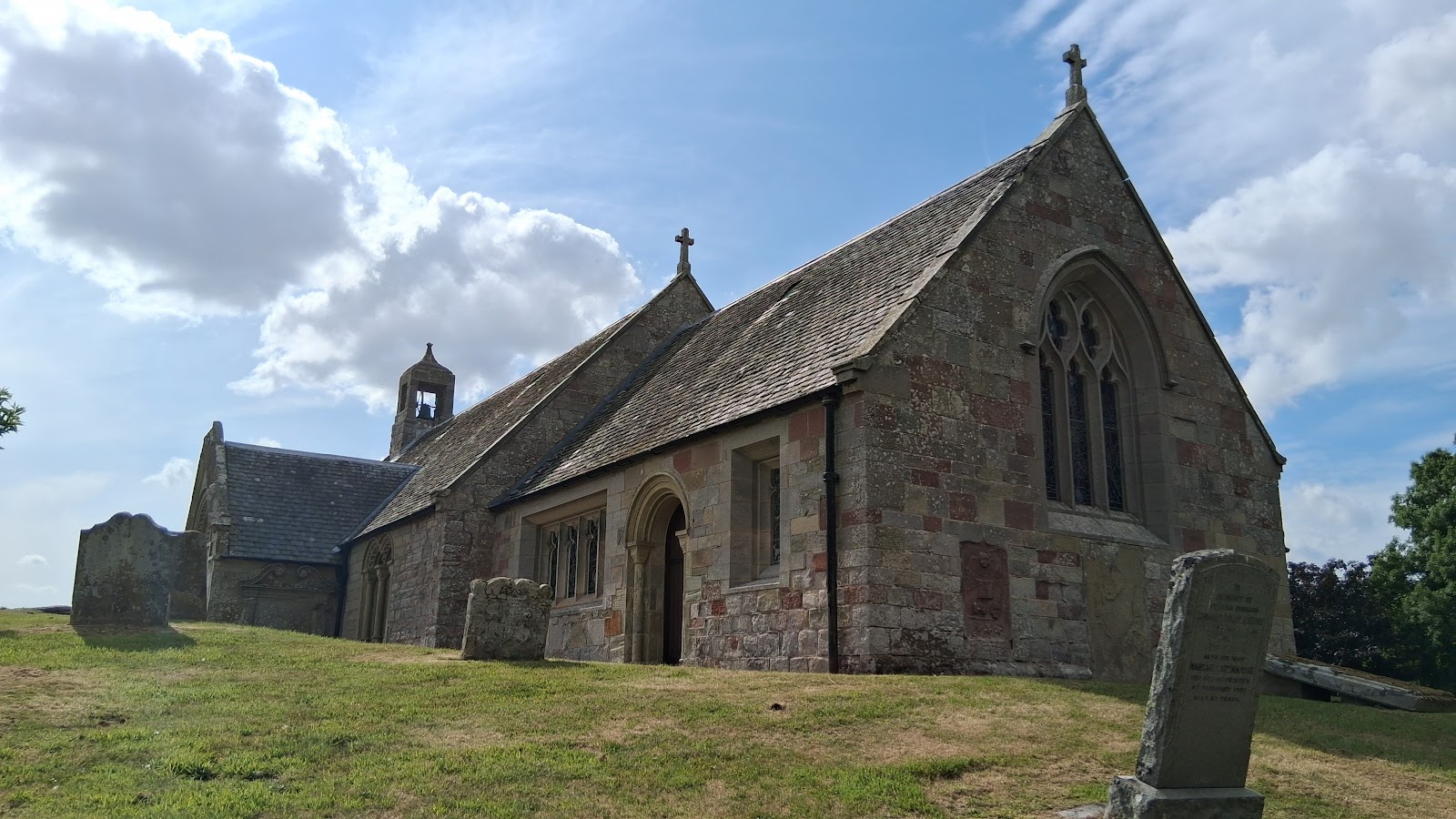 Linton Kirk on its grassy knoll, a stone church with a small bellcote, among old gravestones under a summer sky.